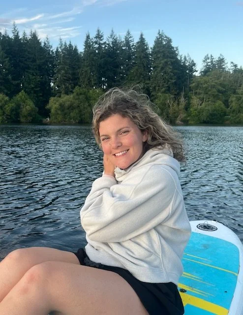 A young woman with curly hair sitting on a paddleboard on a lake, smiling and resting her face on her hand, surrounded by trees.
