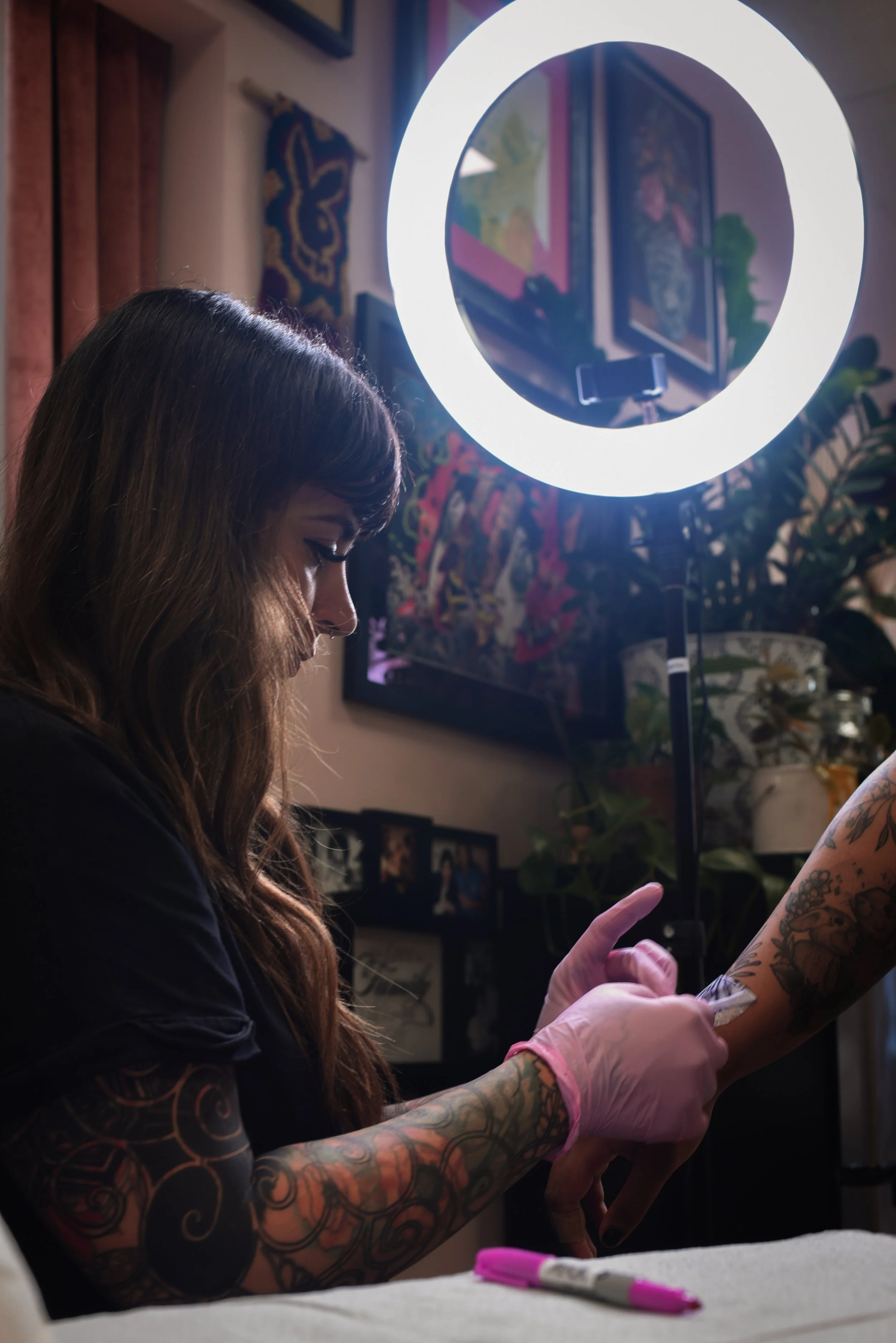 A woman with tattoos and pink gloves getting a tattoo on her forearm, illuminated by a ring light, in a room decorated with artwork and photos.