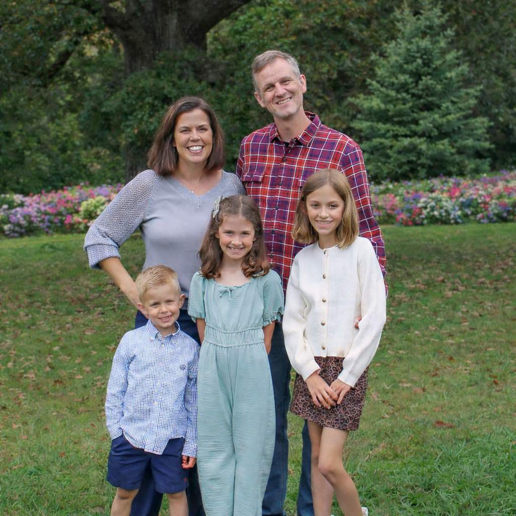 Family of five outdoors in a garden with trees and flowers, smiling for the camera.