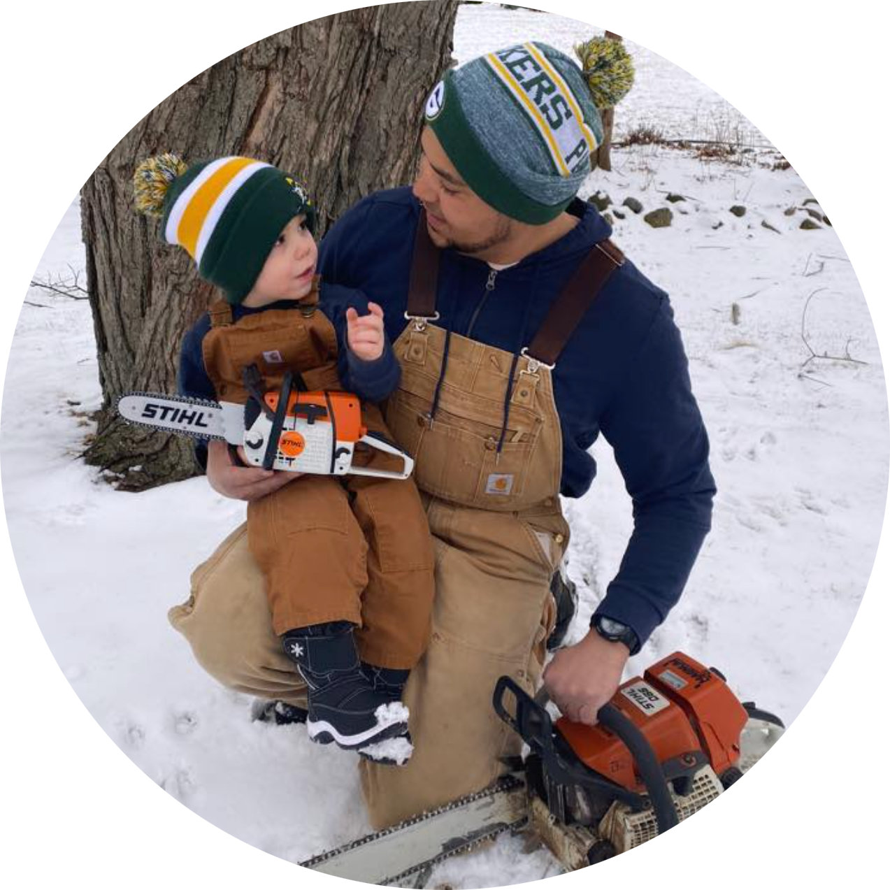 A man in beige coveralls kneeling in snow, holding a young child dressed in winter clothing and a hat, while holding a chainsaw, outdoors near a tree.