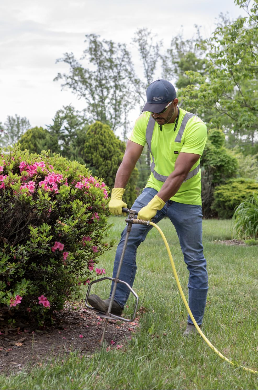 A man wearing a neon yellow shirt, jeans, and yellow gloves using a gardening tool to remove a bush with pink flowers in a garden.