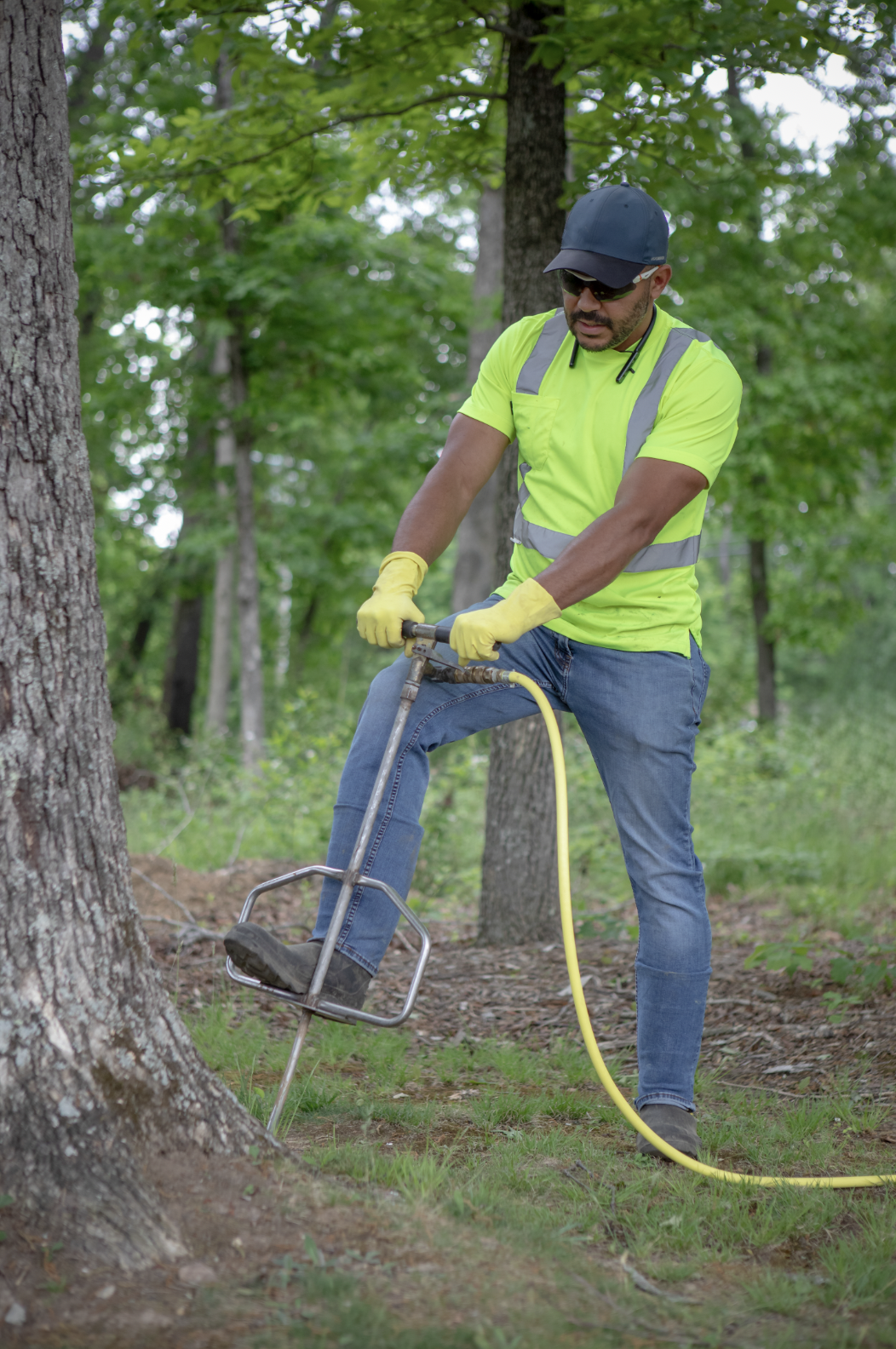 A man wearing a yellow safety vest, yellow gloves, sunglasses, and a blue cap using a power auger to drill into the ground near a tree in a wooded area.