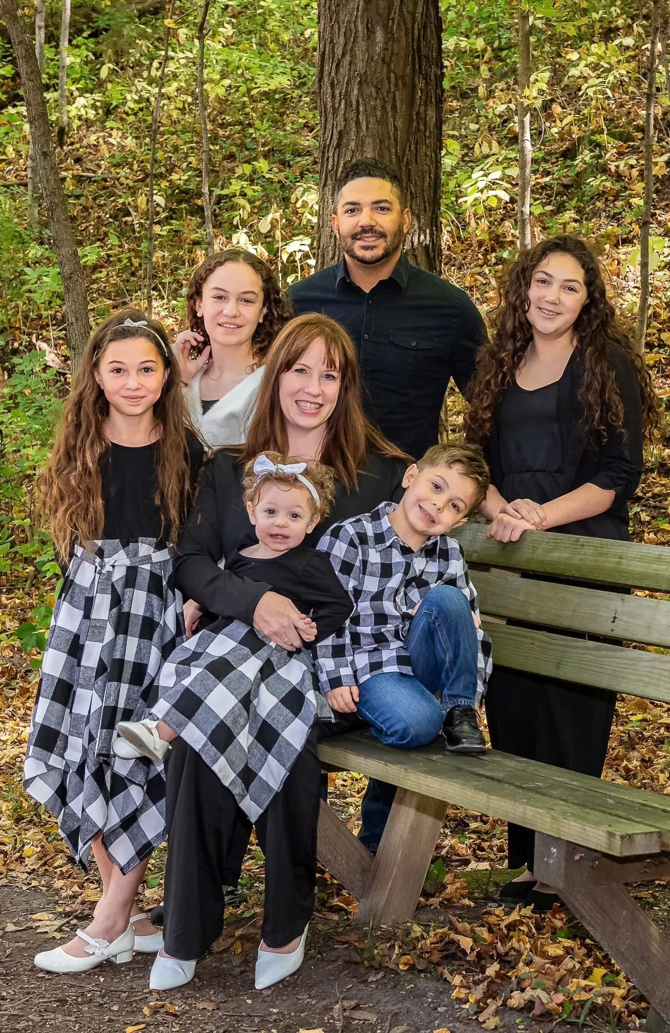Family of seven posing outdoors in a forest, standing and sitting on a wooden bench, with autumn foliage.