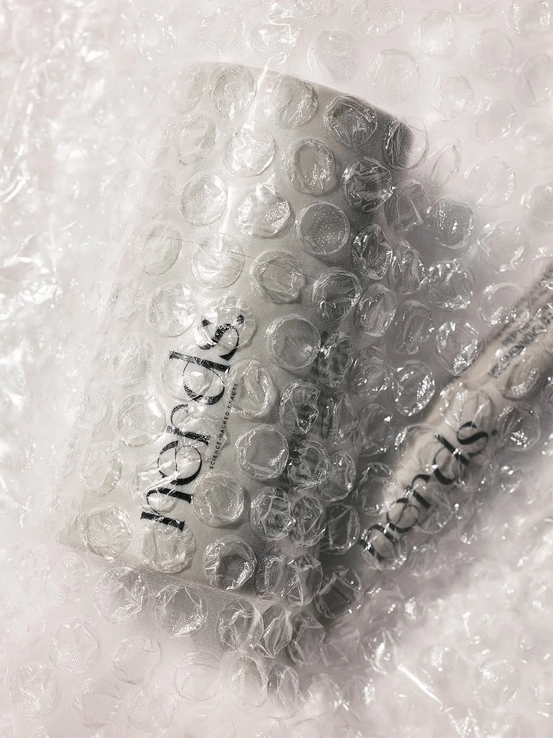 nerds. skincare bottle and tube pressed into clear bubble wrap on crumpled white paper, soft diffused light, tactile editorial beauty product photography