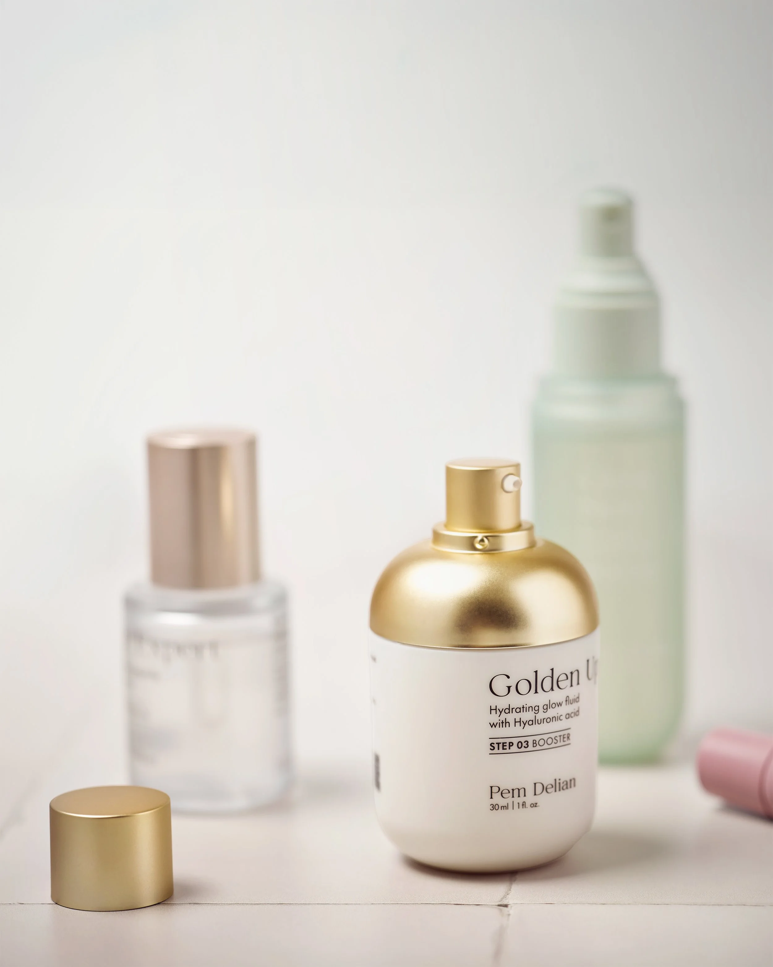 Pem Delian skincare routine lifestyle shot: Golden Up booster, Age Expert serum, and Glass Skin toner on marble surface with canvas pouch and pearl accessories