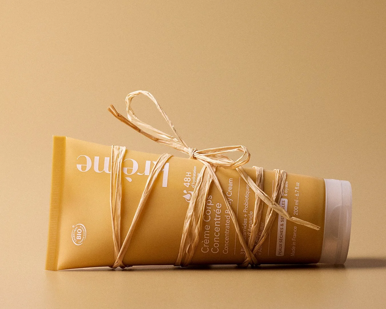 Krème Concentrated Body Cream tube lying horizontally, wrapped in natural raffia string tied in a bow on warm golden background