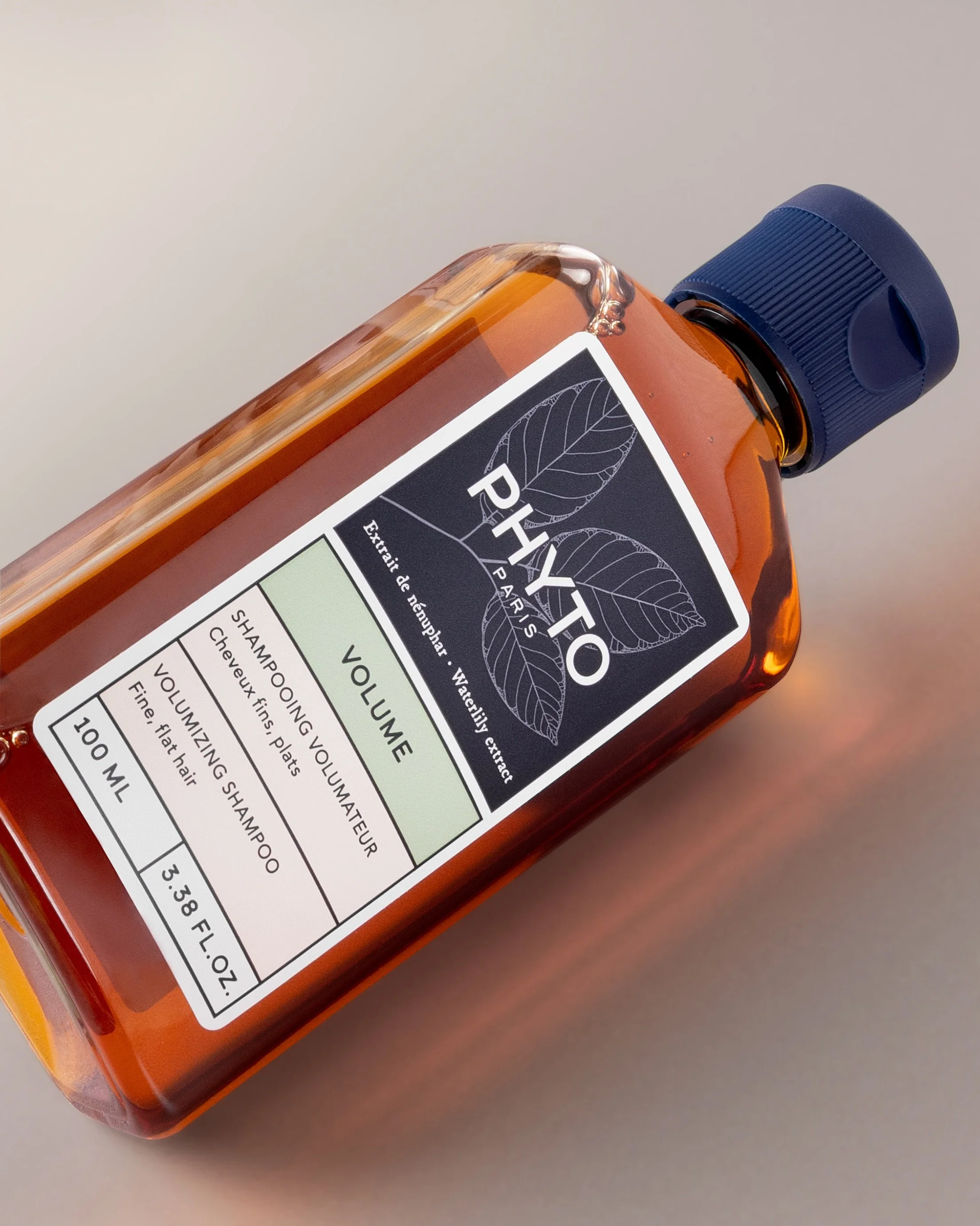 Close-up of an amber glass shampoo bottle placed on its side on a neutral background. The minimalist label features soft green and cream colour blocks, while warm light passes through the liquid, creating subtle reflections and a calm, premium feel.