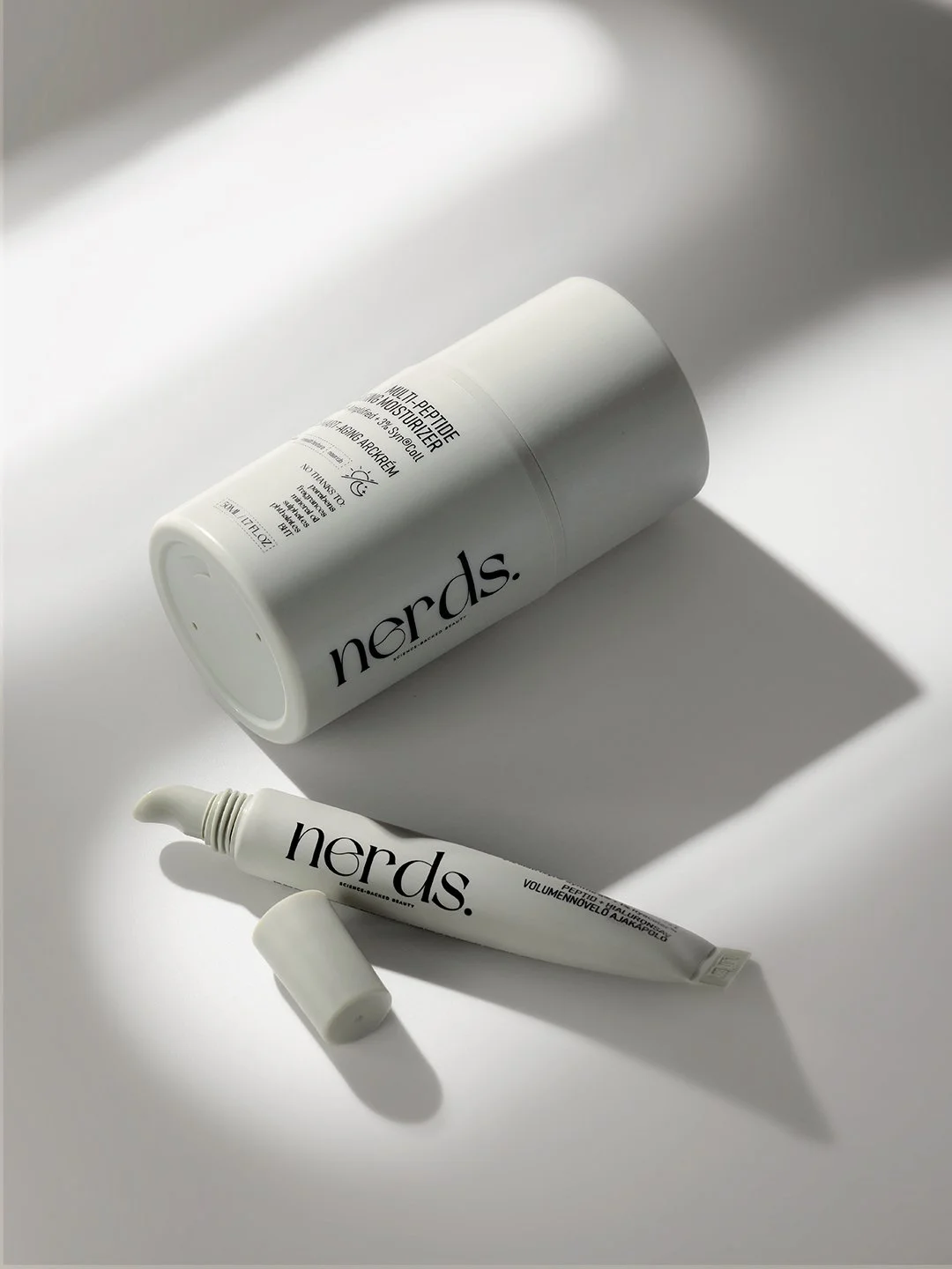 nerds. multi-peptide moisturizer bottle and tube on a white surface, hard directional window light casting long diagonal shadows, skincare product photography