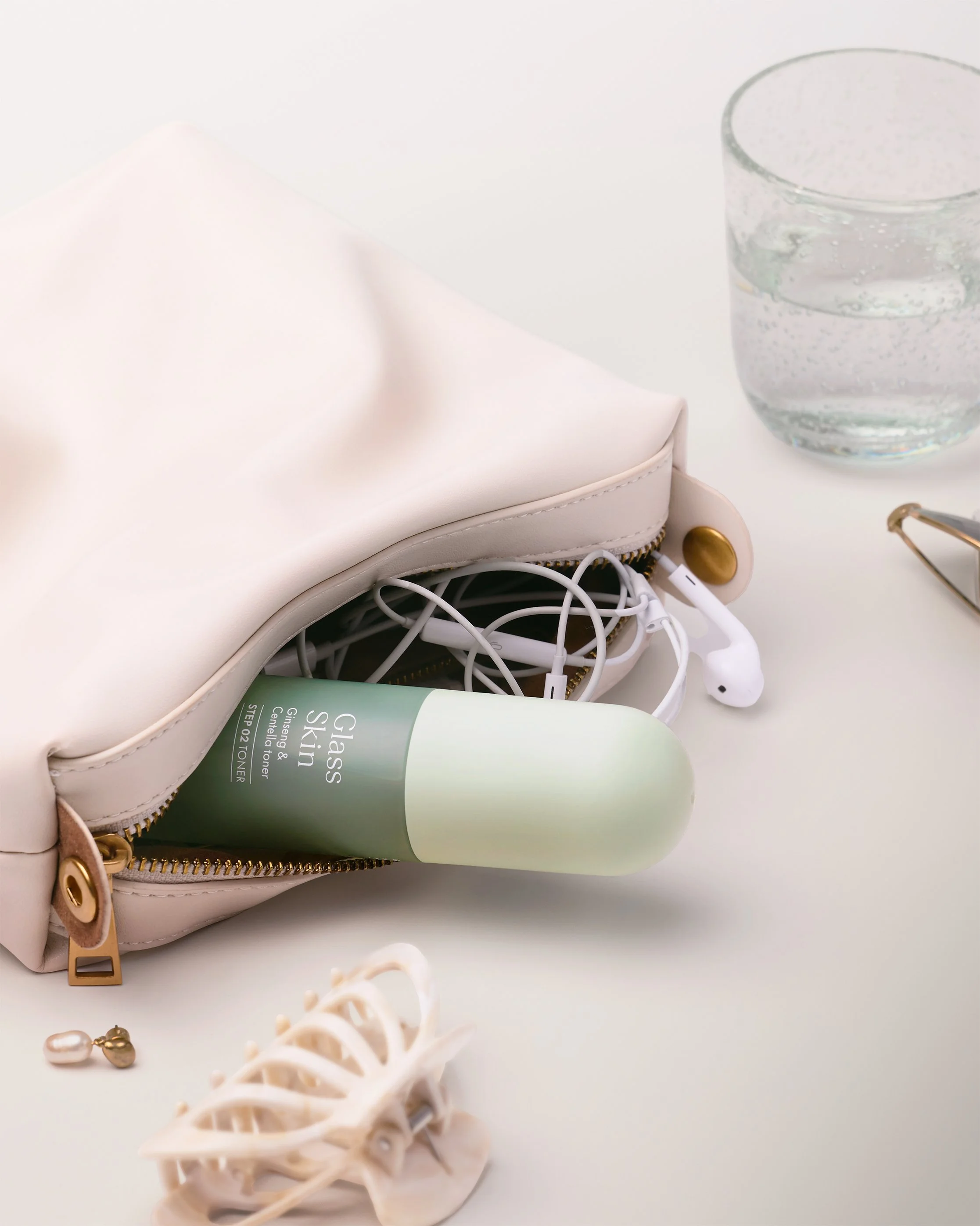 Pem Delian Glass Skin Ginseng & Centella toner in cream leather pouch with wired earphones and pearl earrings on white surface