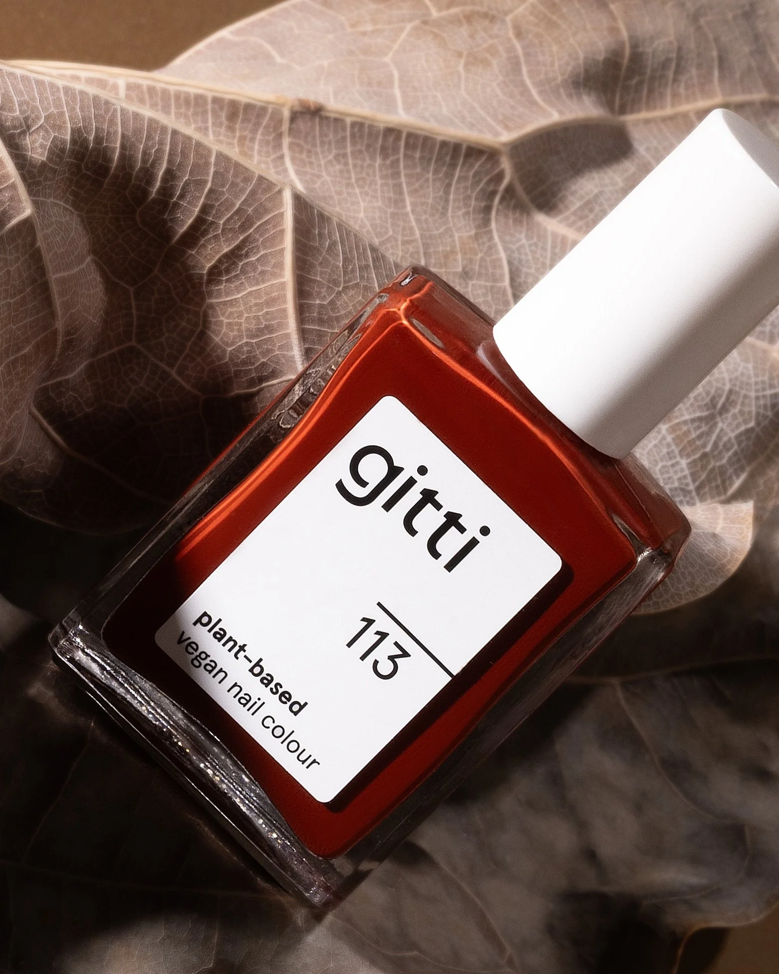 Close-up editorial product photo of Gitti plant-based nail polish shade 113 on dried leaf texture. Warm shadows, minimal styling, clean beauty branding.