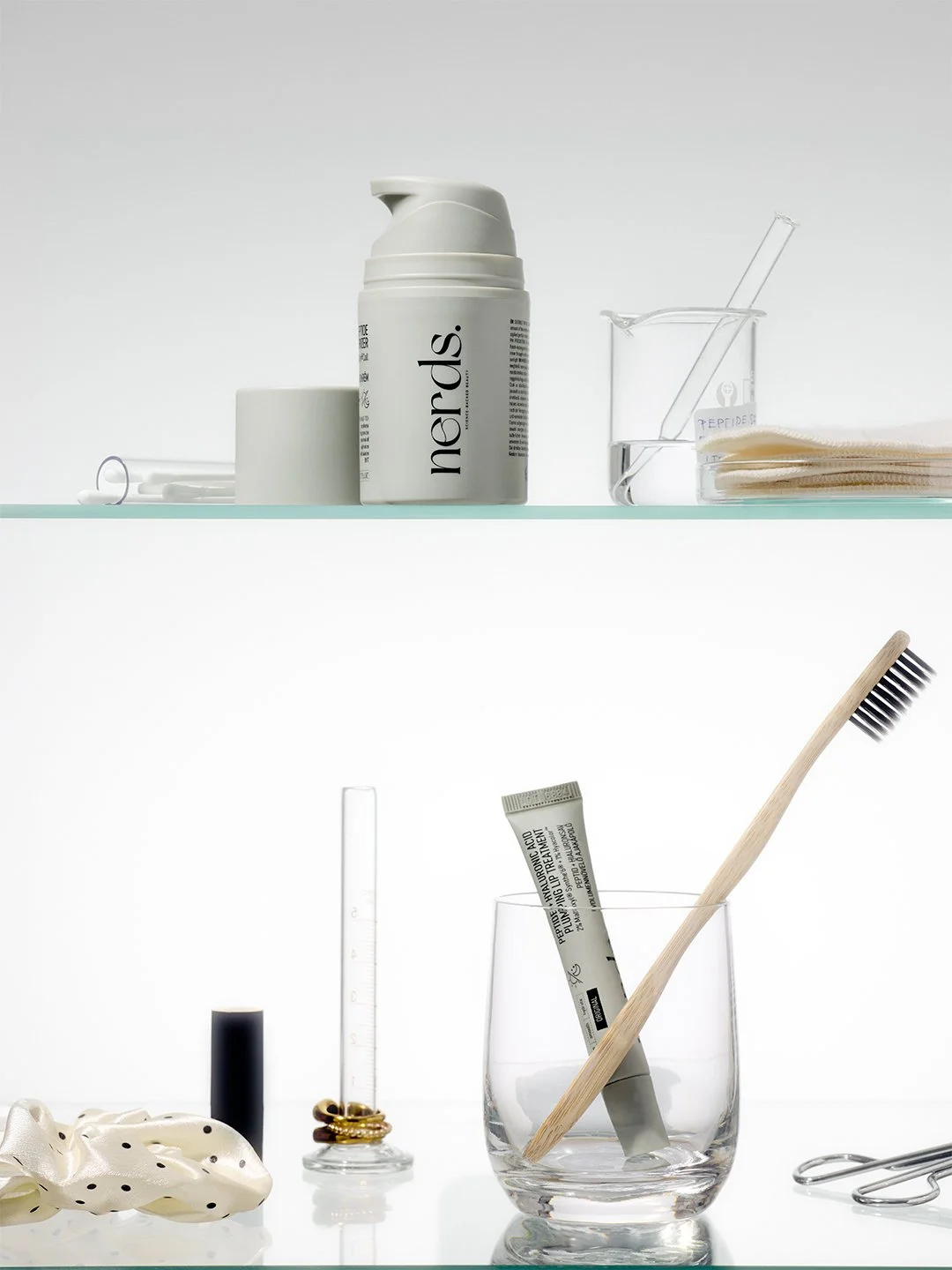 nerds. clinical skincare product photography, pump bottle and tube styled on a glass bathroom shelf with a lab beaker, bamboo toothbrush and gold rings in soft white light
