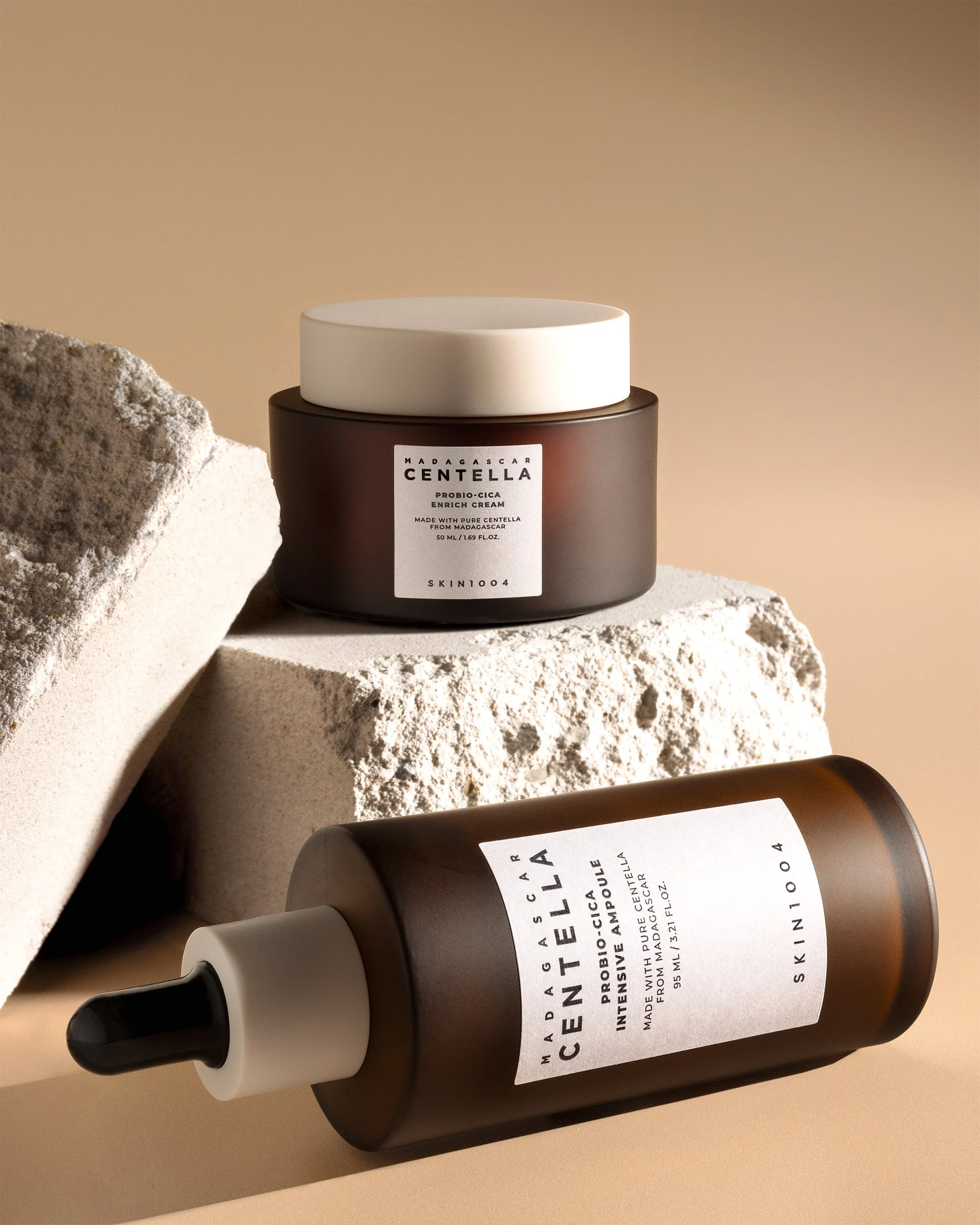 Skincare jar and brown ampoule bottle placed on a textured beige stone surface, lit with soft directional light creating deep shadows and a minimalist still-life composition.