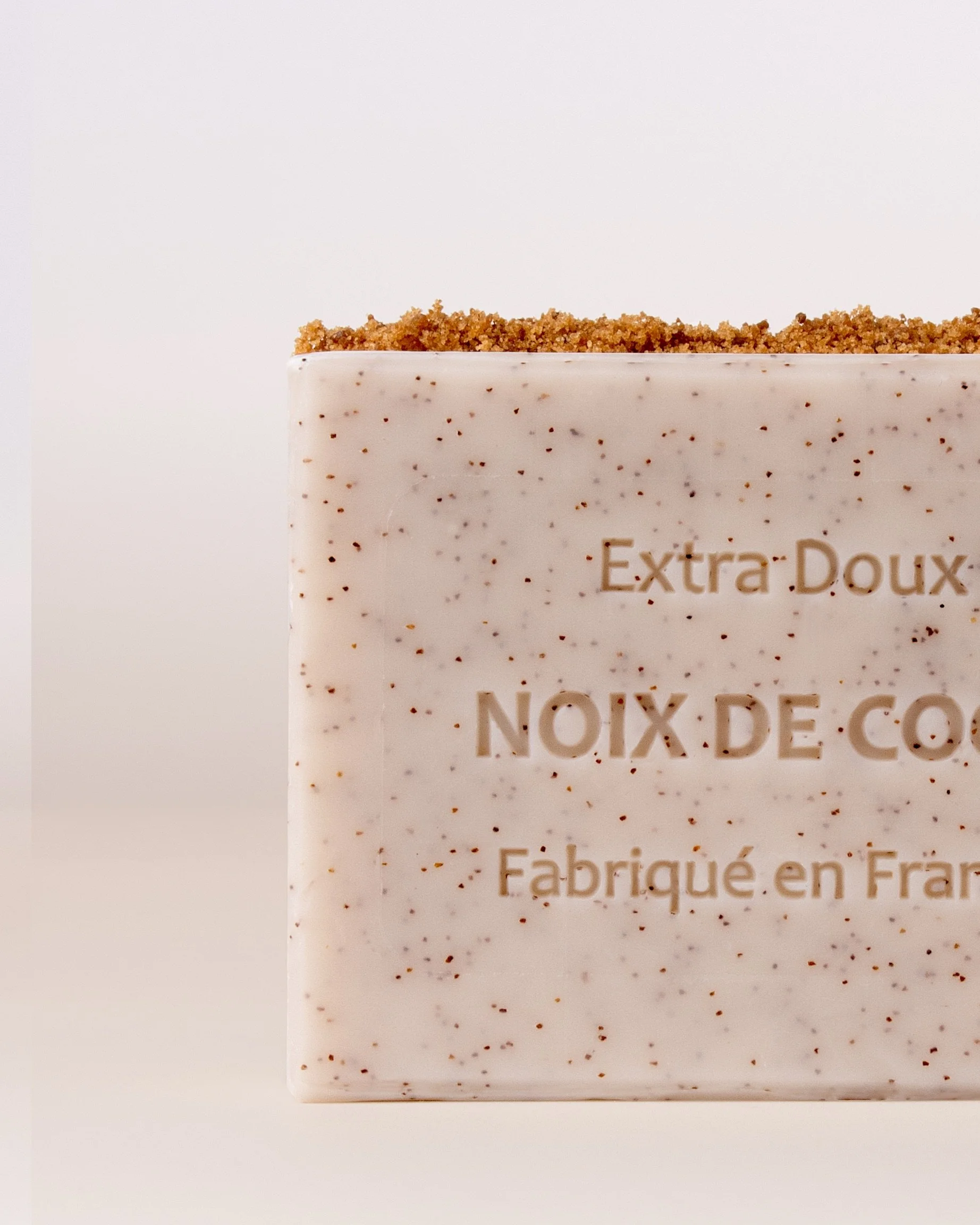 Close-up product photography of a coconut exfoliating soap bar with visible natural speckles and a textured crumb layer on top, featuring embossed French text, photographed against a soft neutral background with natural light.