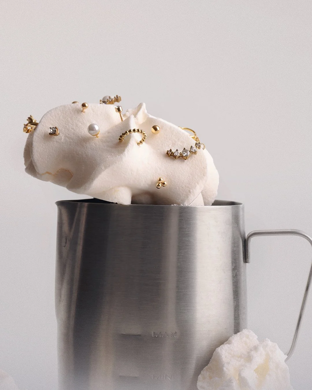 conceptual, prop-led jewelry photography. Stack of cracked meringue domes holding an assortment of gold rings, studs, a pearl, and pavé pieces, arranged emerging from a stainless steel milk pitcher