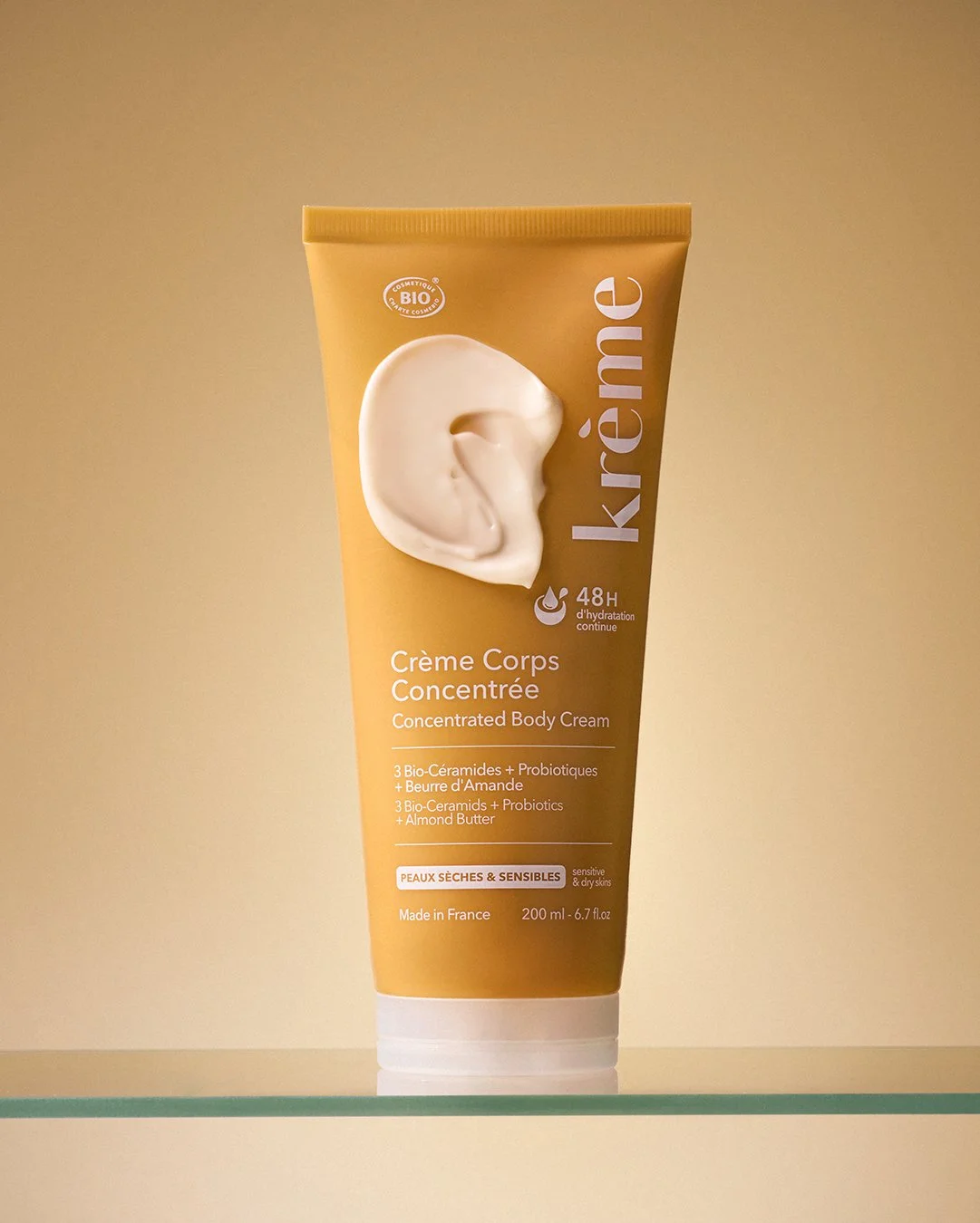 Krème Crème Corps Concentrée standing upright on a glass surface with a generous cream swatch spread across the front of the golden tube. Rich, silky texture front and centre — minimal styling, maximum impact. Made in France. PureCapture Media. 📍 Ha