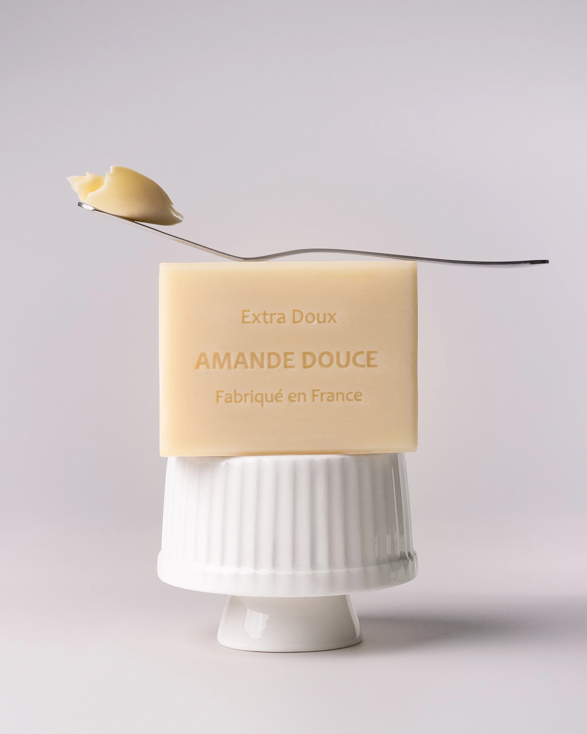 Minimal product photography of a French almond soap bar stacked on a white ceramic pedestal, with a stainless steel spoon holding a creamy butter texture above the product, styled against a soft neutral background with natural light.