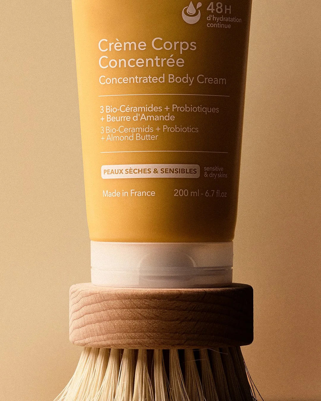 Close-up of Krème Concentrated Body Cream tube balanced on a round wooden body brush — natural bristles meet bio-ceramide skincare. Earthy texture, warm tones, French organic beauty photography. For sensitive & dry skin. PureCapture Media. 📍 Haarlem