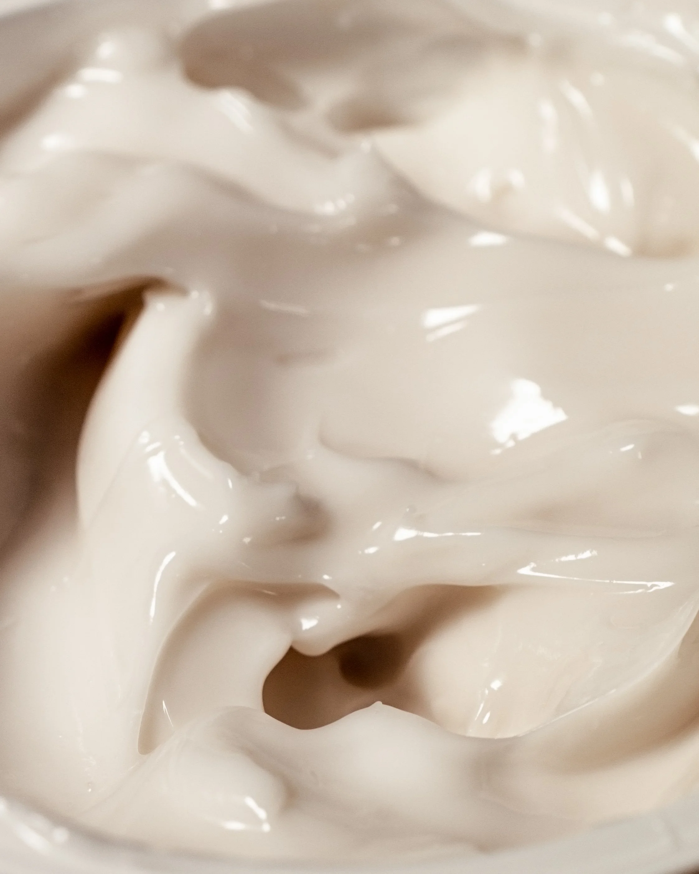Close-up macro photograph of a rich white skincare cream with soft peaks and glossy highlights, showing texture and density.