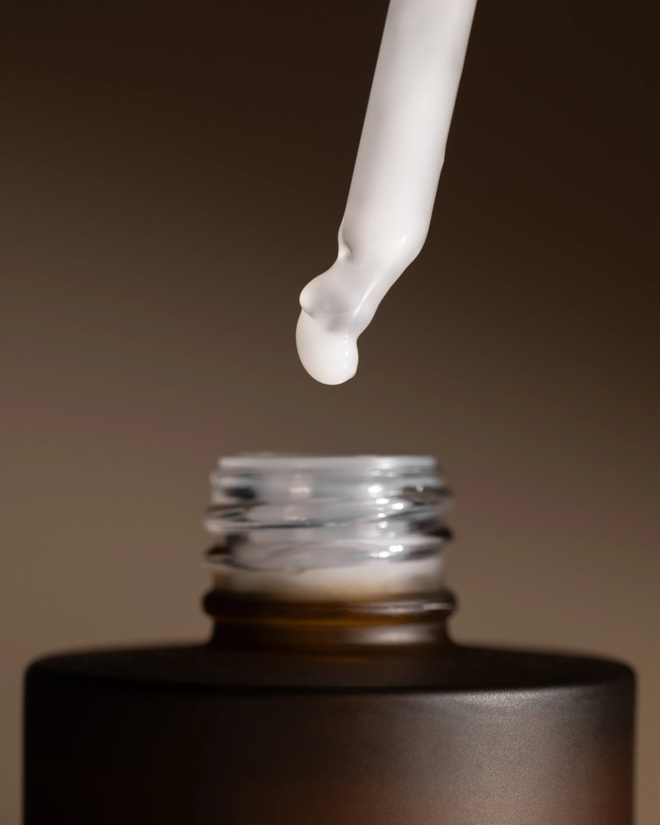 Glass dropper releasing a serum drop into the opening of a brown skincare bottle against a warm gradient background.
