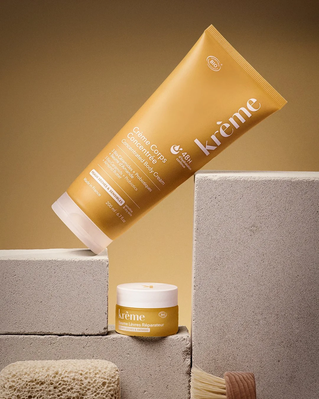 Krème body cream tube leaning diagonally against concrete blocks with lip balm jar, pumice stone and brush in foreground