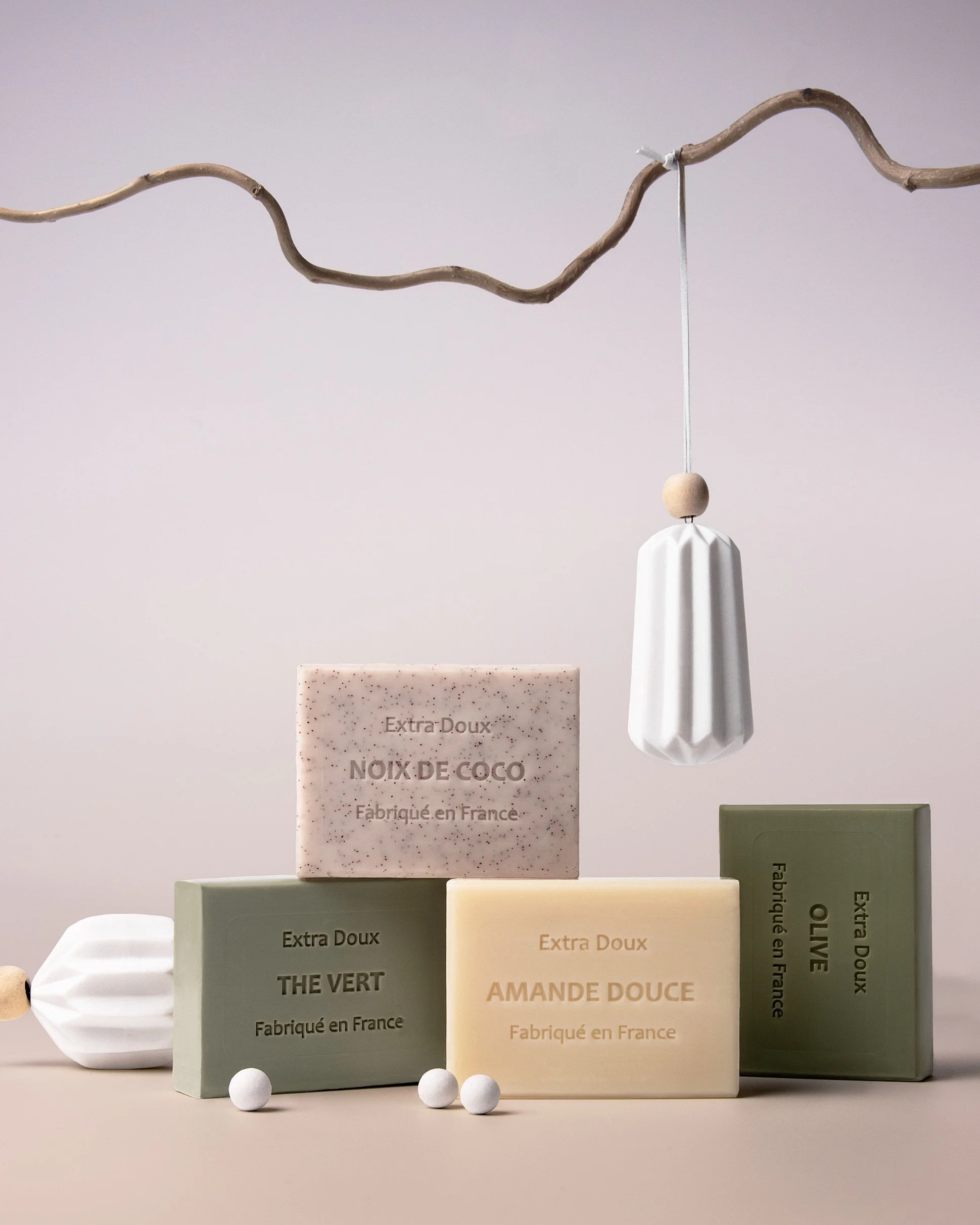 Stacked French soap bars in muted green, cream, and beige tones styled with a ceramic hanging ornament and a curved bare branch. Minimal holiday still-life with modern apothecary aesthetics on a soft beige background.