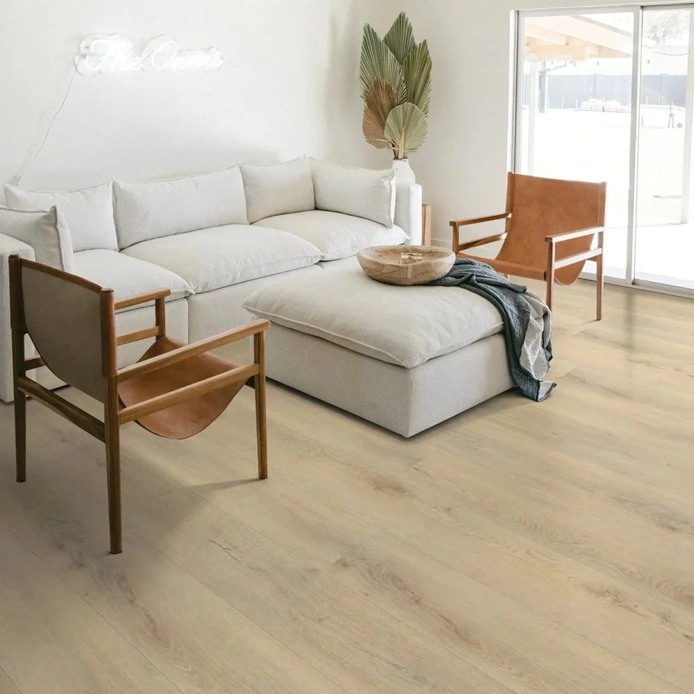 A living room with a white sofa, two wooden chairs with tan leather seats, a wooden bowl, a throw blanket, and a tall vase with large dried palm leaves. There's a neon sign on the wall and sliding glass doors letting in natural light.