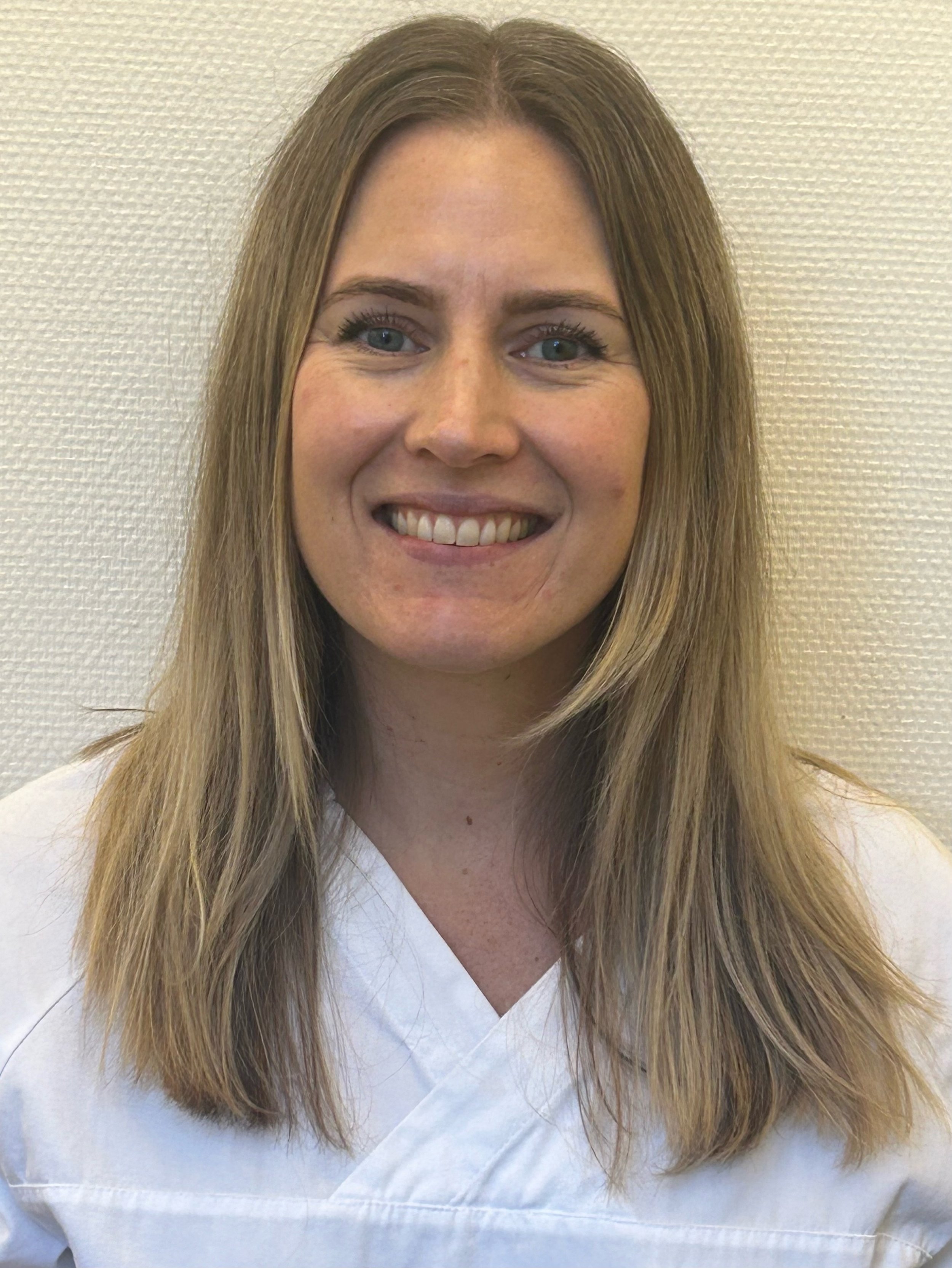 A woman with long, light brown hair and blue eyes smiling, wearing a white top, standing against a light textured wall.
