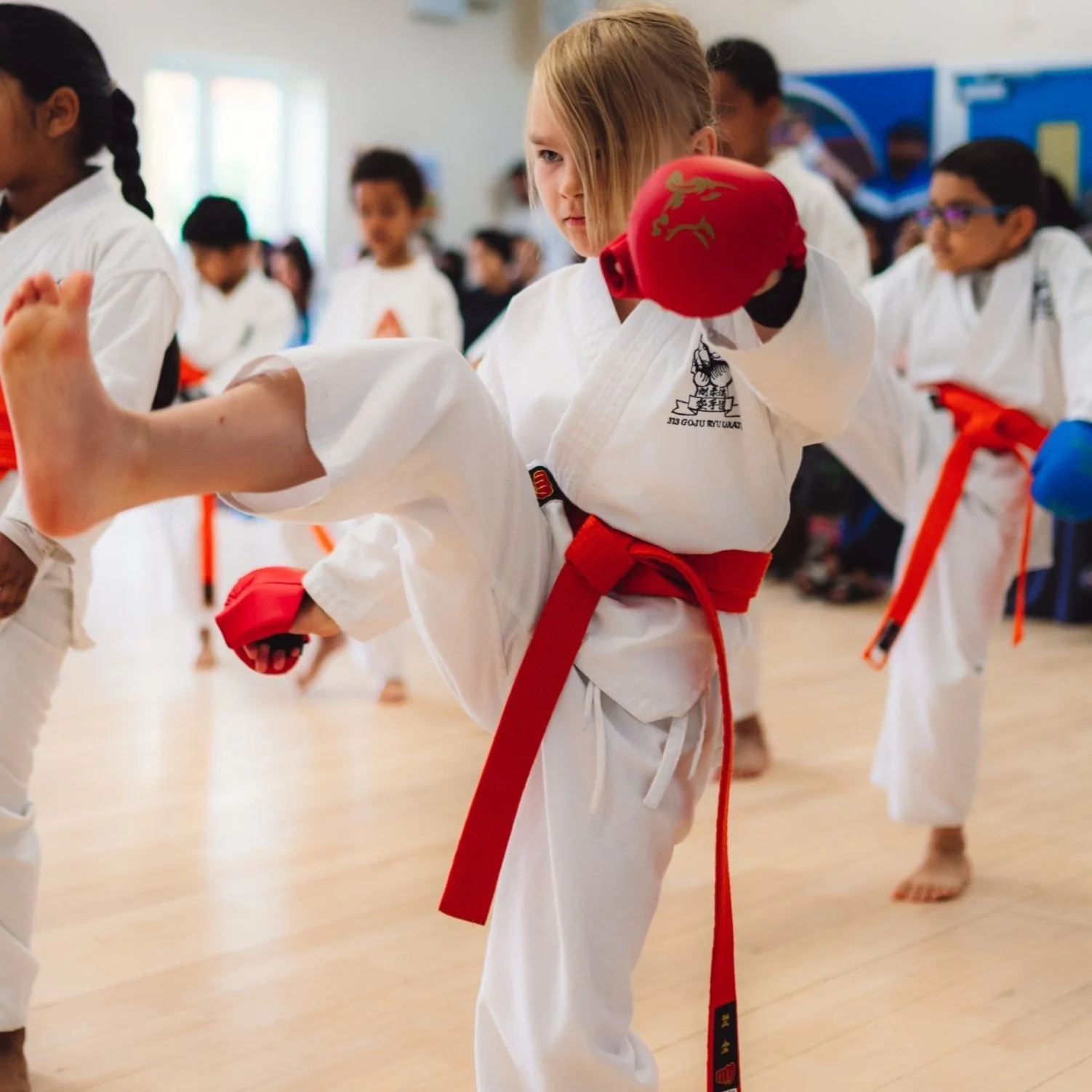 Children practicing karate in a dojo, wearing white karate uniforms with colored belts and sparring gloves.