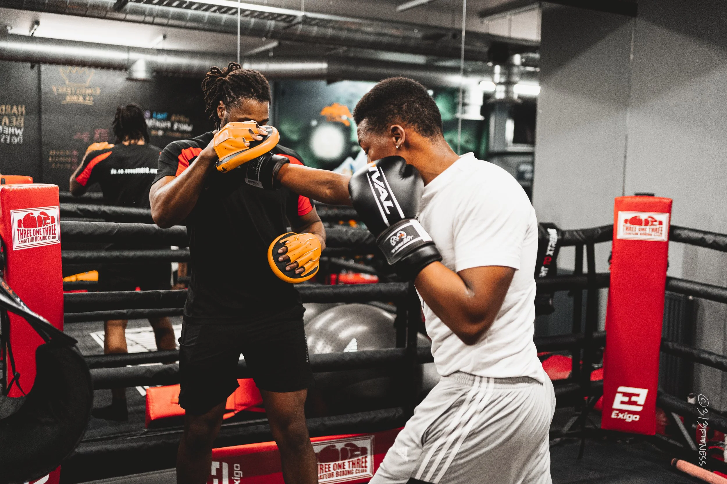 Two men practicing boxing in a boxing gym, with one throwing a punch and the other blocking, surrounded by a boxing ring and trainers.
