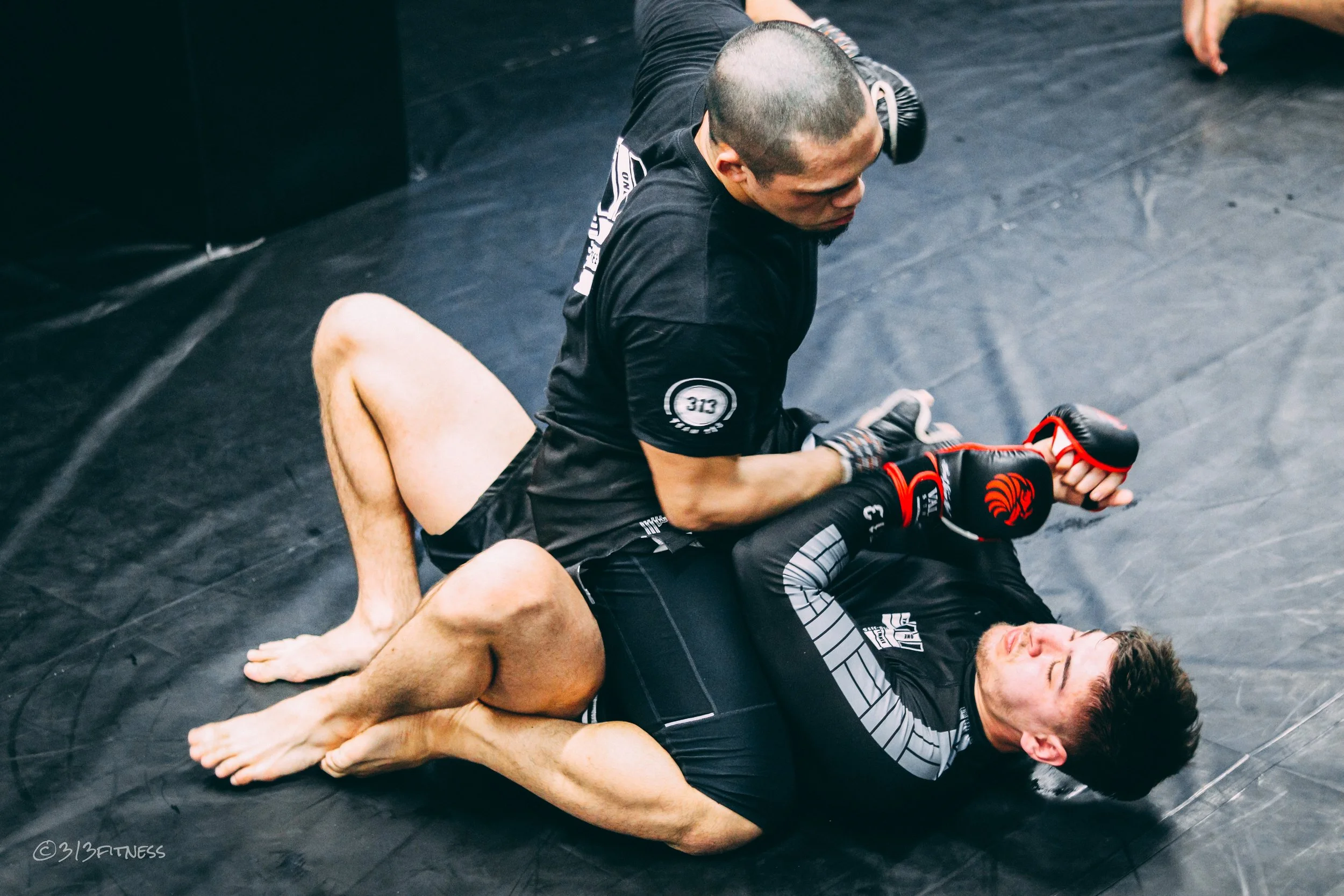 Two men engaged in mixed martial arts training on a black mat; one man is on his back wearing black rash guards and shorts, while the other man is on top wearing black clothing and gloves, about to land a punch.