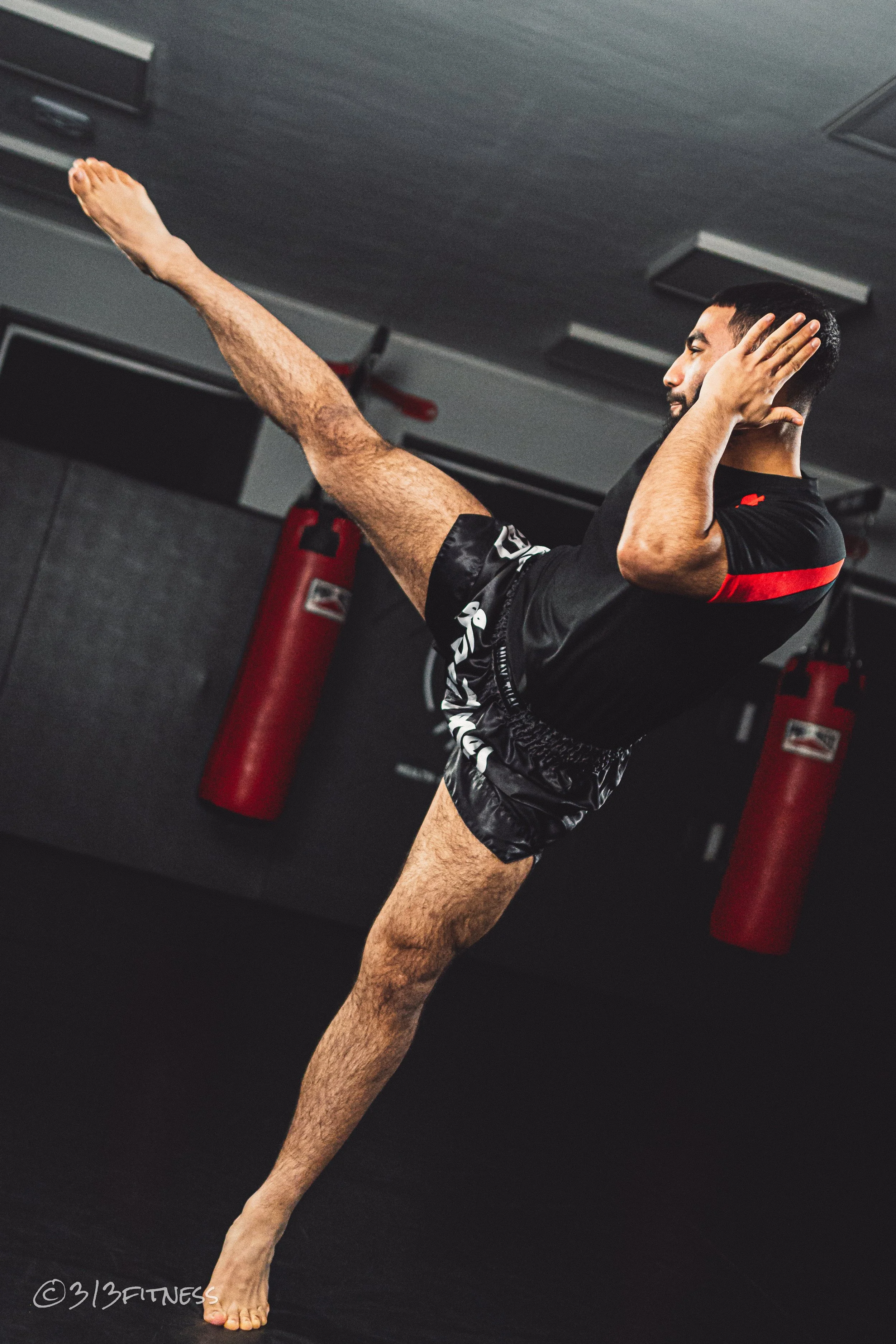 A man practicing martial arts or kickboxing in a gym, performing a high kick with a focus on balance and technique, with punching bags in the background.