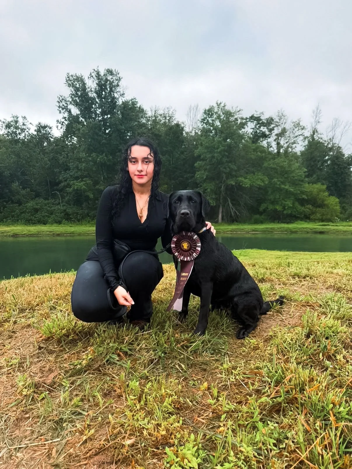A woman with black curly hair and a black outfit kneels next to a black service dog wearing a large award ribbon around its neck, by a pond with trees in the background.