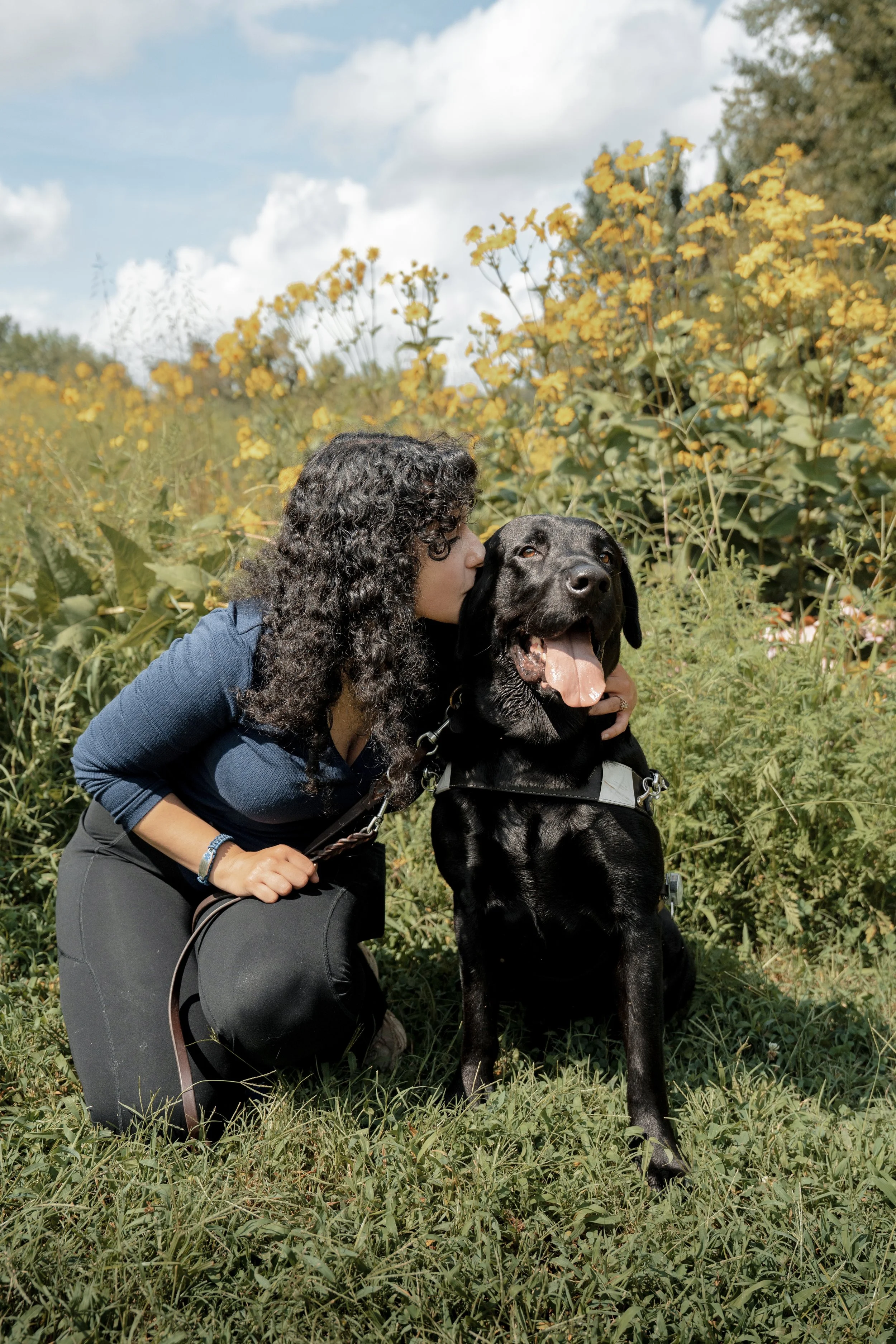 A woman with curly hair kissing a black Labrador retriever in a field with yellow flowers and blue sky.