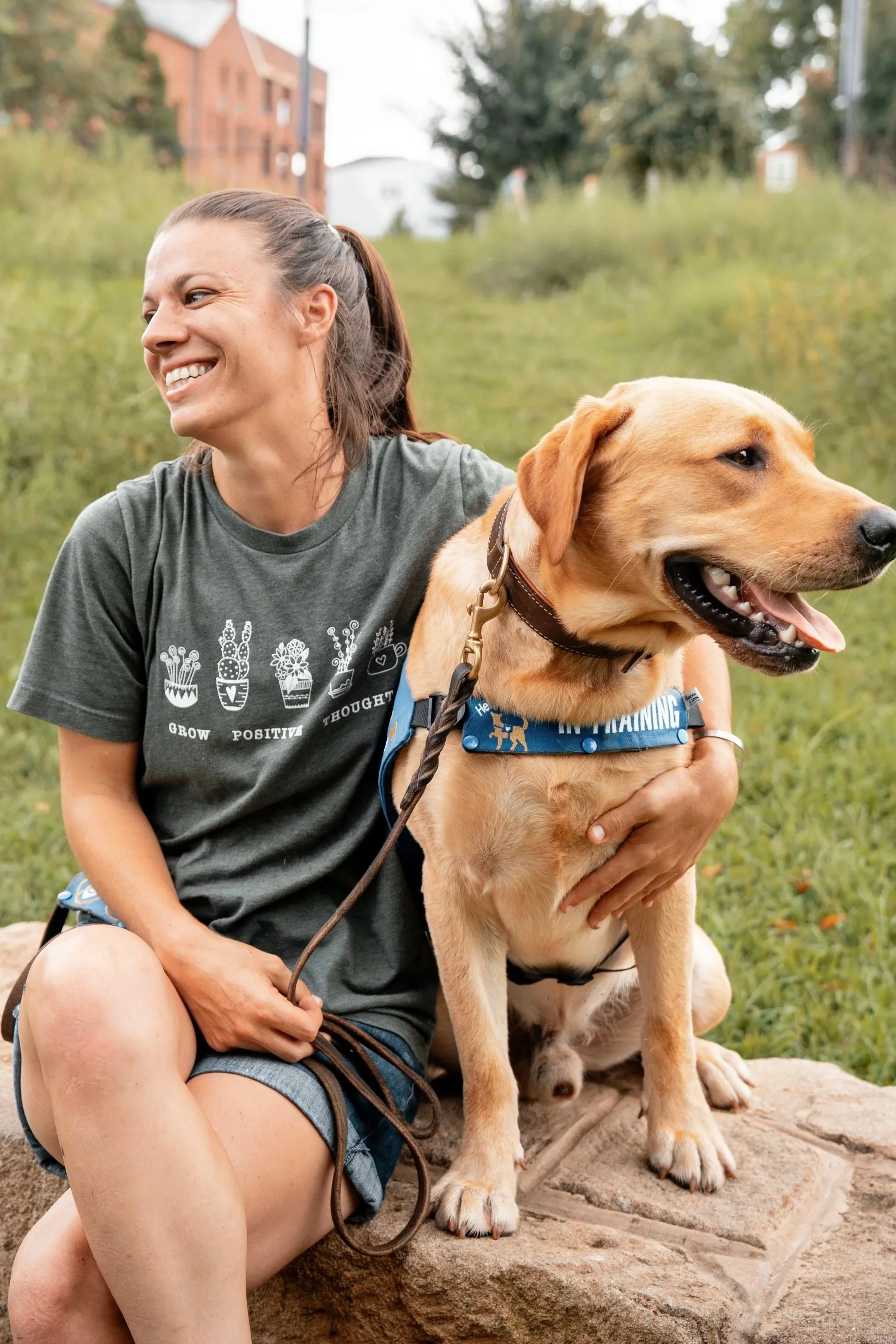 A woman smiling while sitting outdoors on a rock, holding a large yellow Labrador retriever dog on a leash. The dog is wearing a blue vest and sitting beside the woman. The background includes trees and buildings, indicating a park or urban outdoor setting.