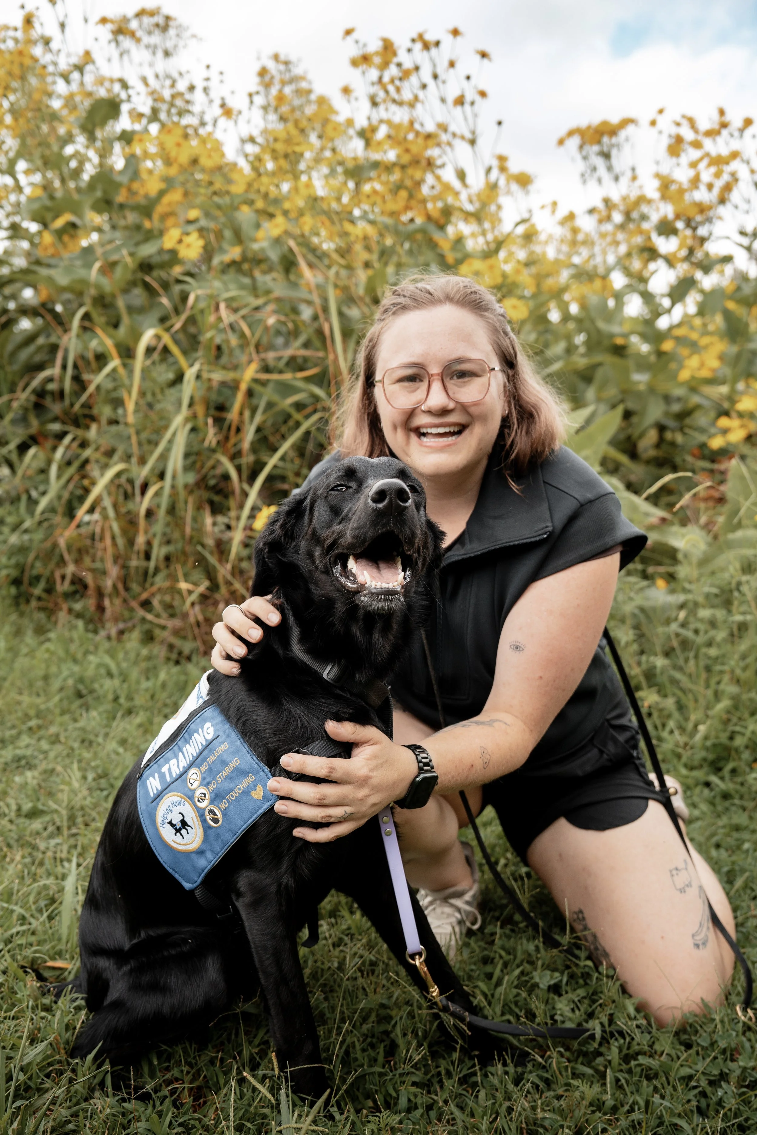 A woman with glasses, tattoos, and brown hair smiling and crouching outdoors with a black dog wearing a service dog vest in front of yellow blooming plants.