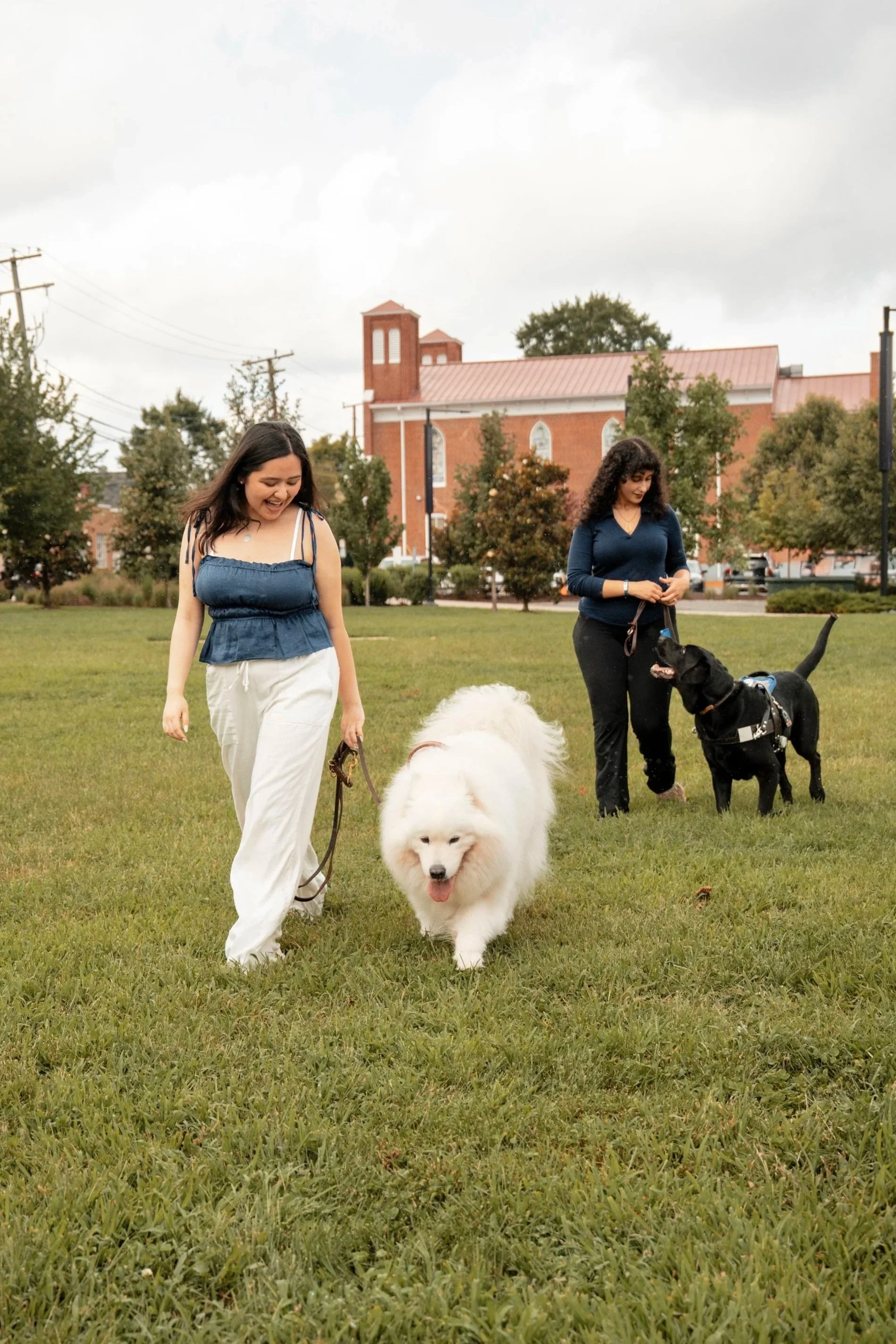 Two women walking dogs in a park with a brick church building in the background.