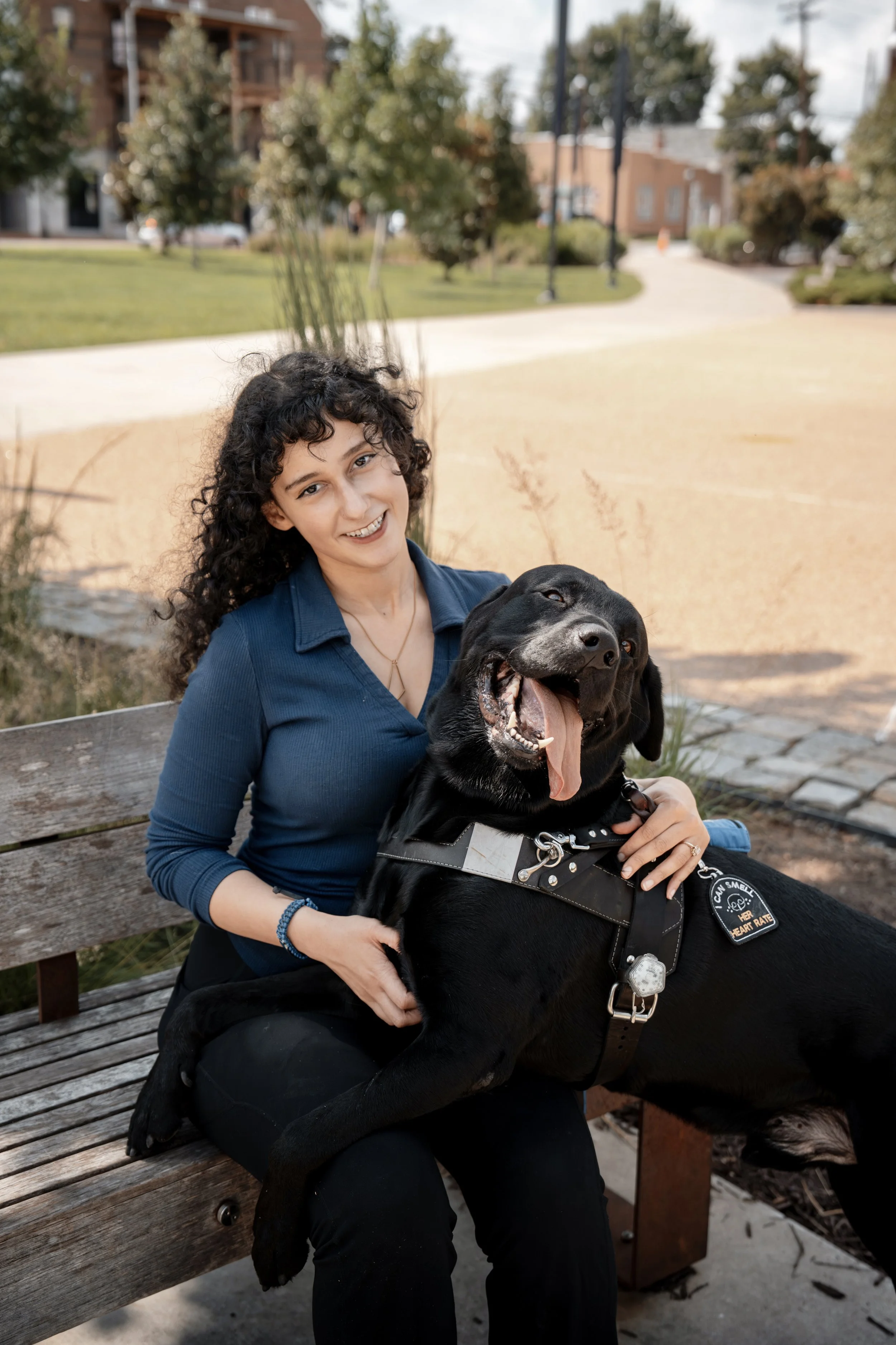 A woman with curly dark hair sitting on a wooden park bench, smiling, with a happy black Labrador Retriever dog wearing a service vest, in an outdoor park with trees, buildings, and a path in the background.