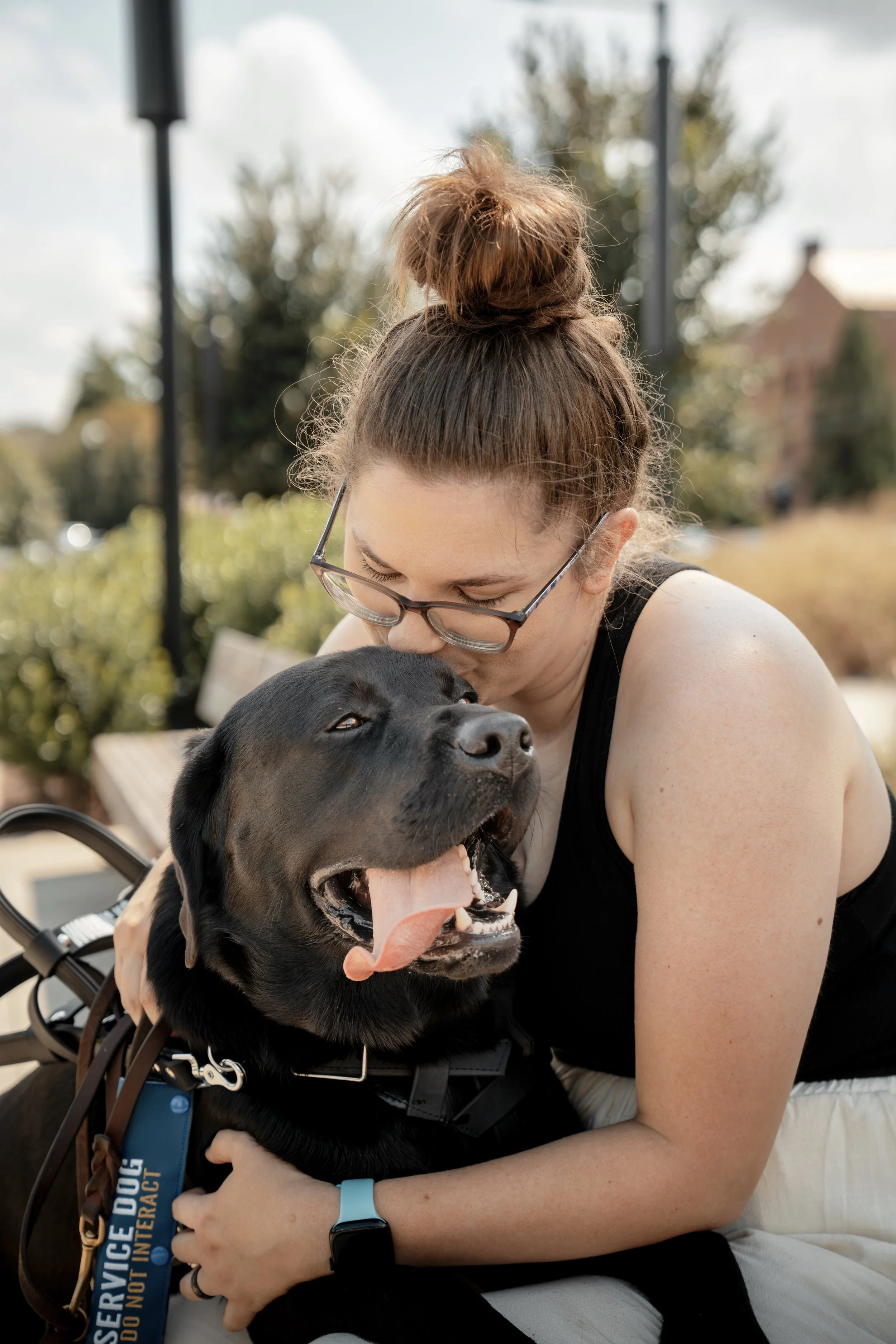 A woman with glasses and a bun hairstyle hugging a large black dog with its tongue out and eyes partially closed outdoors on a sunny day.