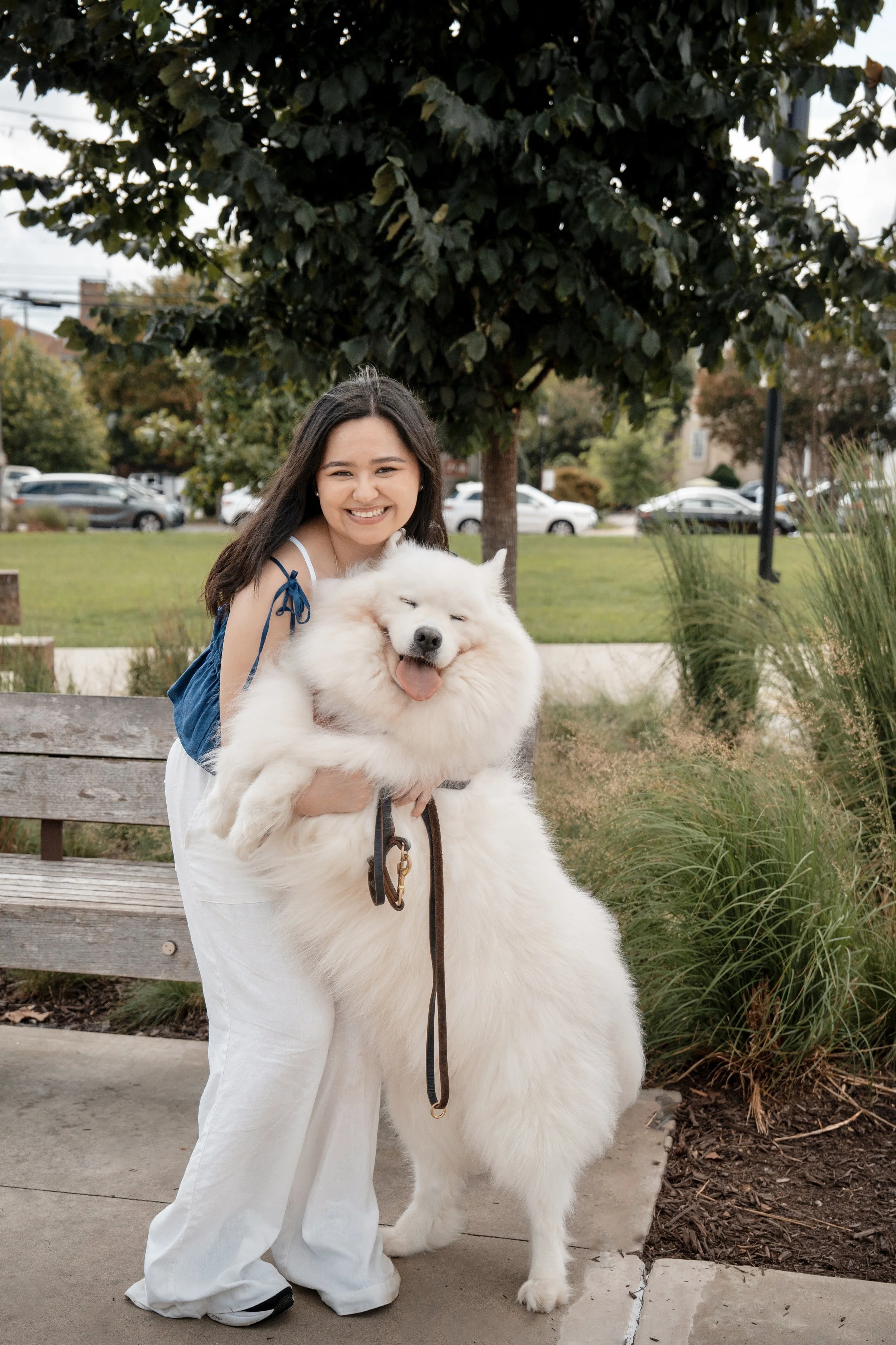 A young woman smiling and holding a large fluffy white dog outdoors in a park or garden setting, with trees and parked cars in the background.