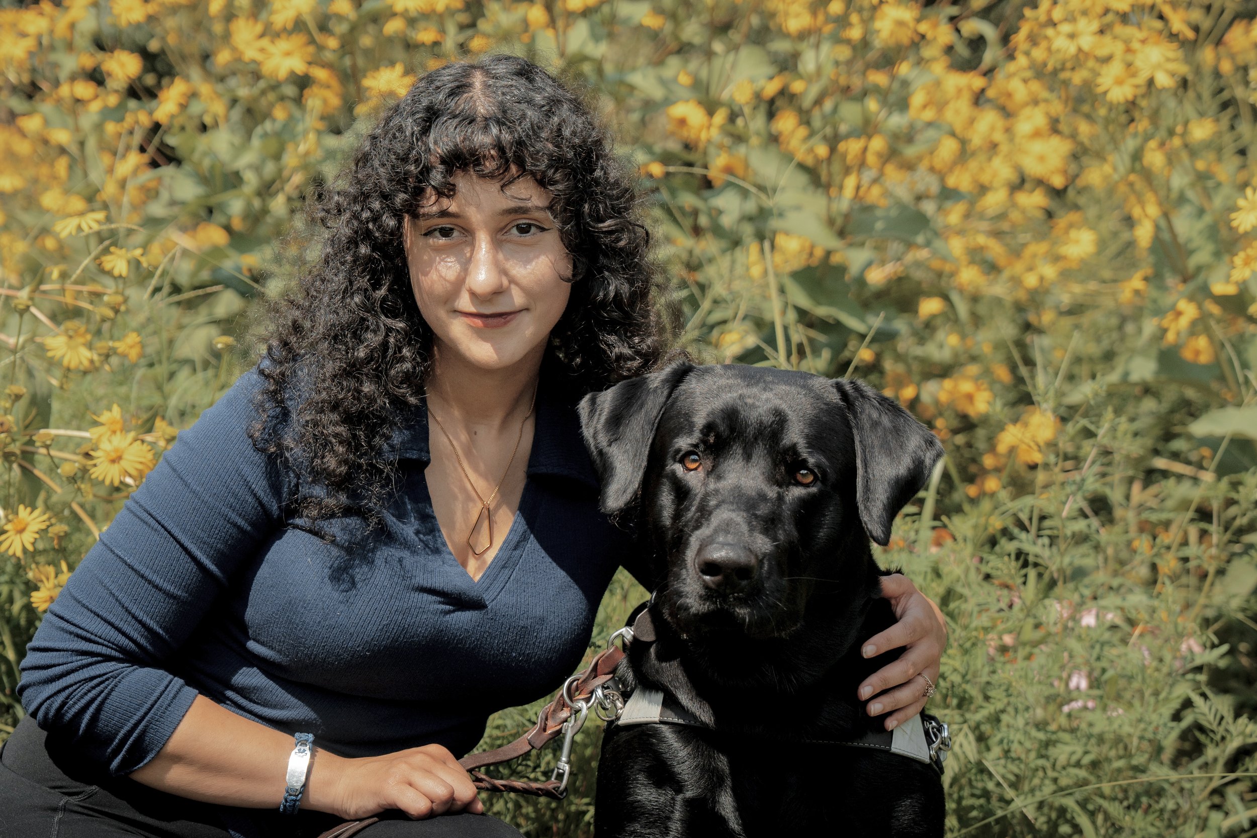 A woman with curly dark hair and a black dog with a chain collar, both outdoors in front of yellow flowers.