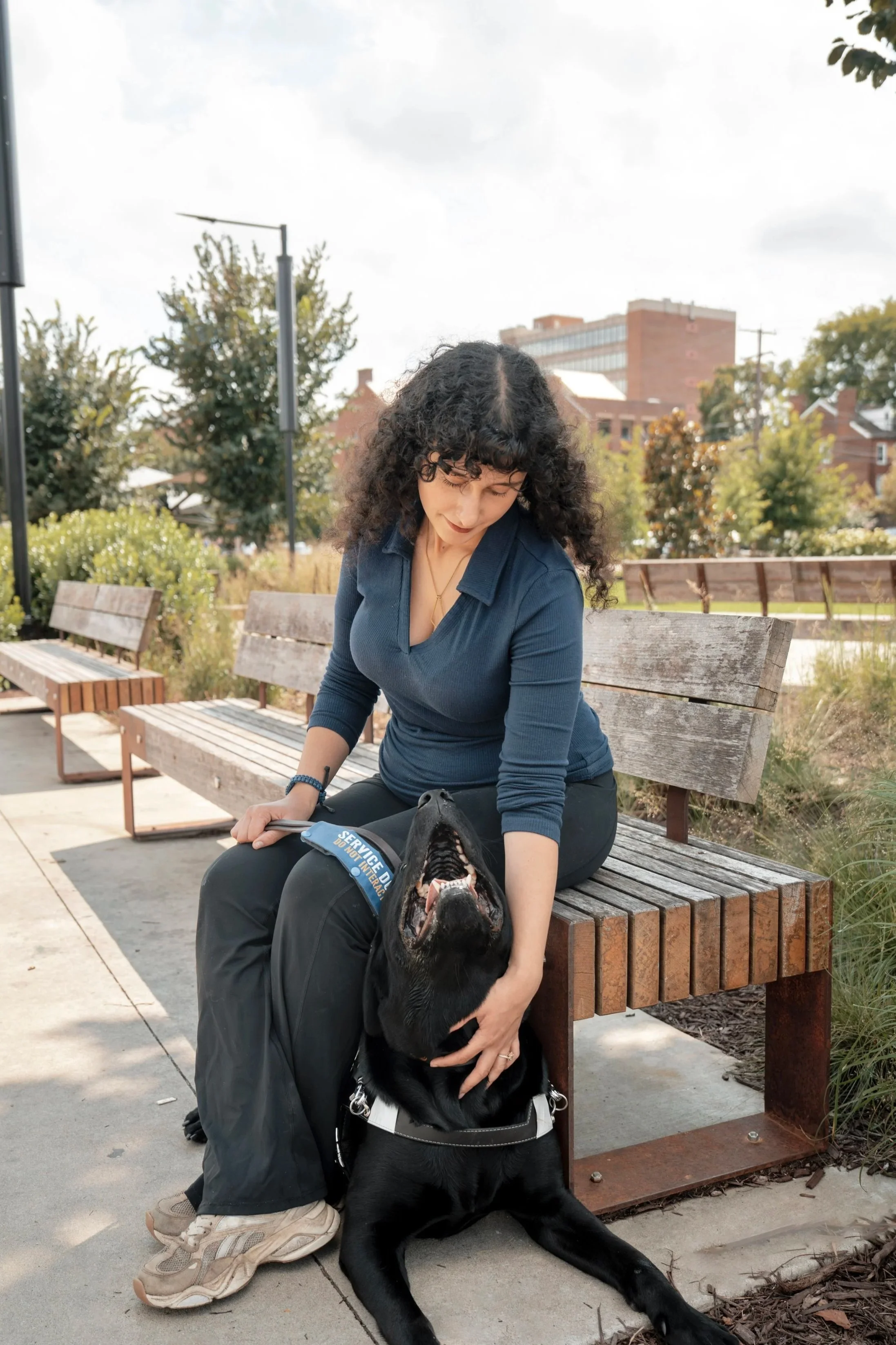A woman with curly hair sitting on a park bench, petting a black police dog lying on the ground with its mouth open. The woman and dog are outdoors in an urban park setting with trees and buildings in the background.