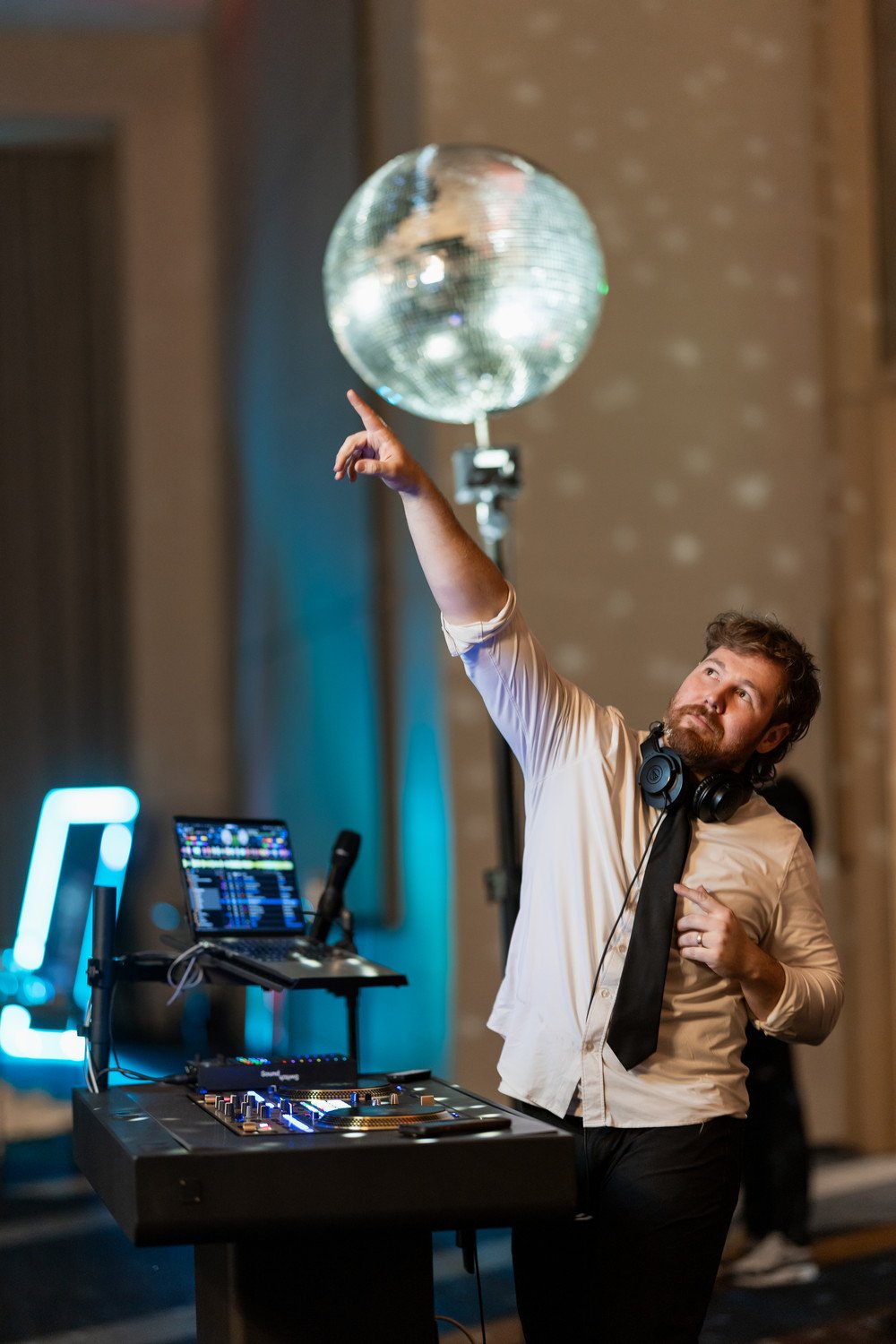 Man in white shirt with black tie and yellow sunglasses DJing at a party, wearing headphones, with a large disco ball and colorful lighting in the background.