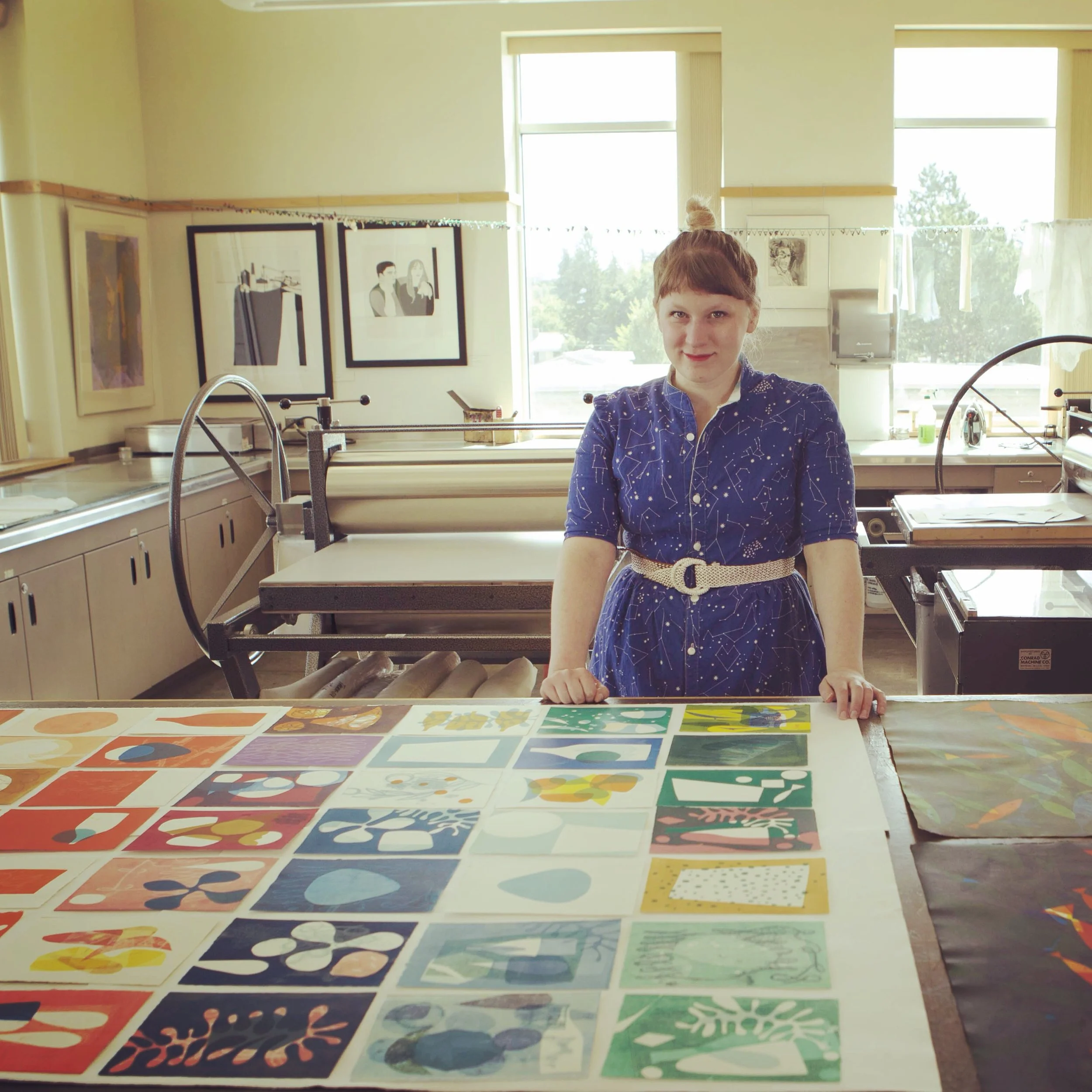 A young girl with red hair and a braid, wearing a blue dress with white constellation patterns, stands behind a table displaying various colorful artworks featuring geometric and abstract designs, inside a well-lit room with large windows and art decorations on the walls.