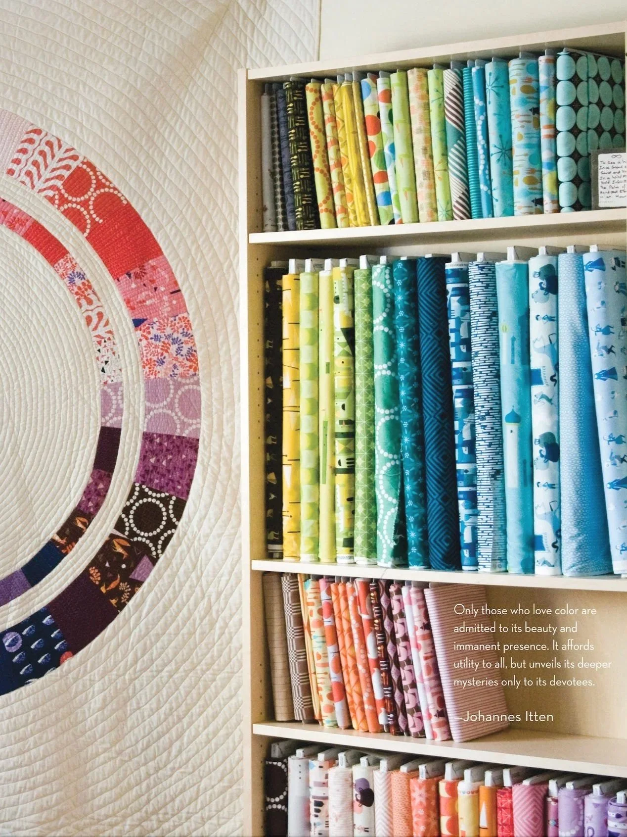 Shelf with neatly folded and arranged colorful fabric bolts in shades of green, blue, yellow, black, and pink, next to a quilt with a circular pattern in red, pink, purple, black, and white.