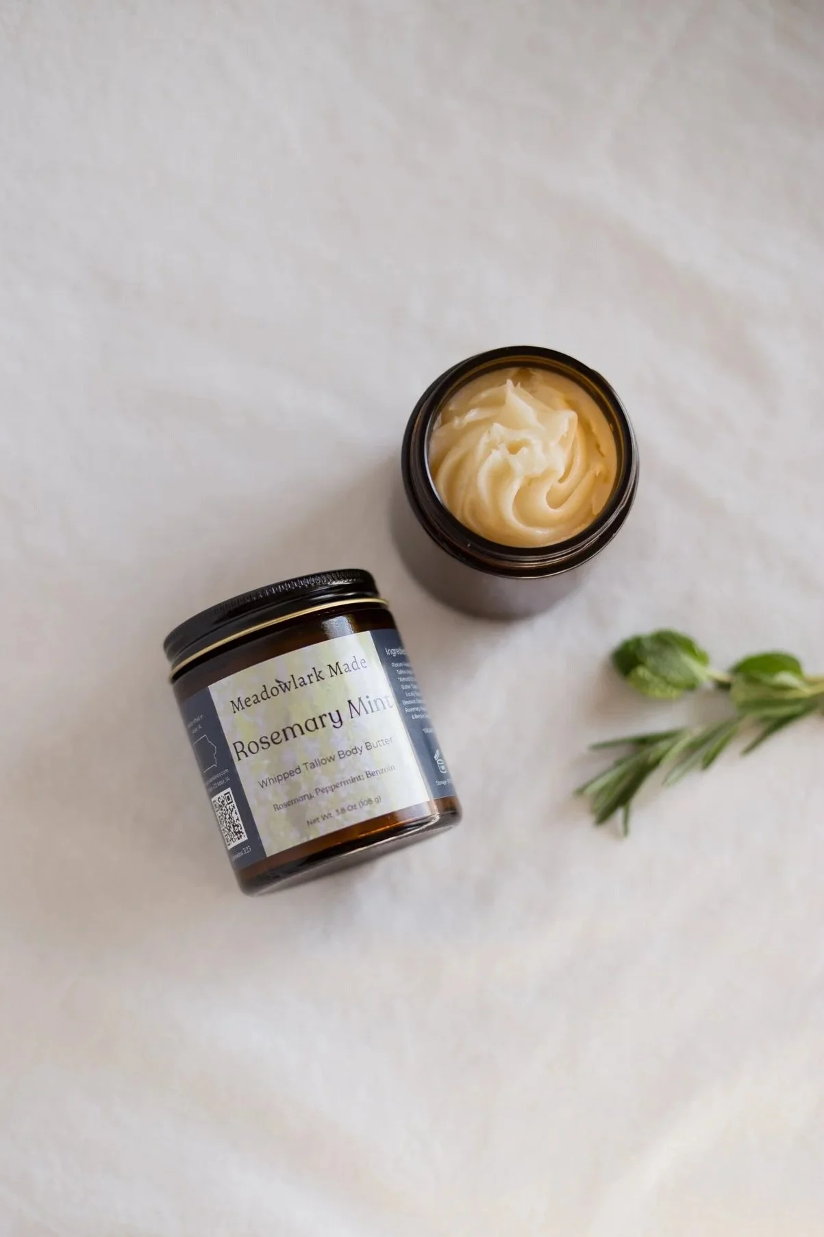 Open jar of Meadowlark Made Rosemary Mint whipped body butter with sprig of rosemary beside it on a white surface.