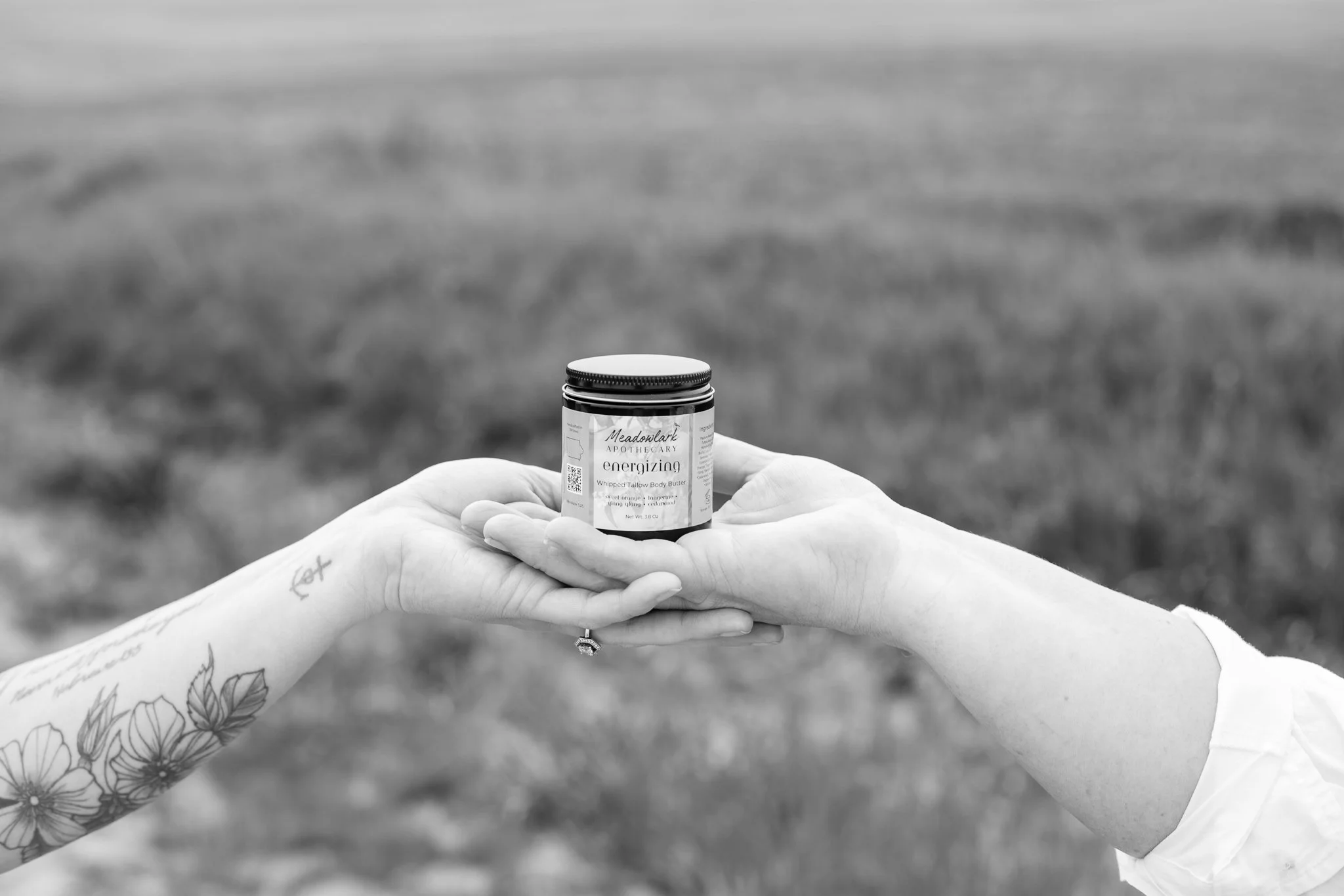 Two generations of hands exchanging a jar of Meadowlark Made: A Prairie Apothecary energizing whipped body butter outdoors, with a grassy field in the background.