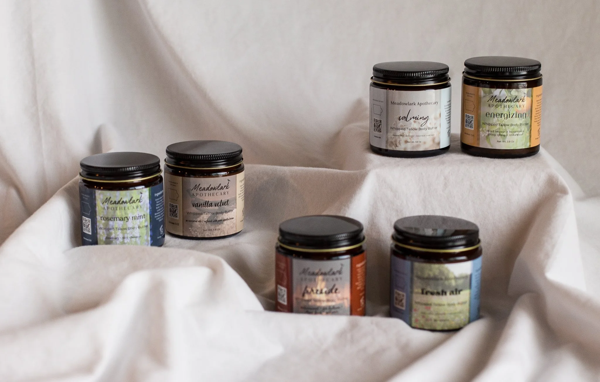 Collection of six jars of whipped tallow body butter with black lids, labeled with different scents and ingredients, displayed on a white fabric surface.