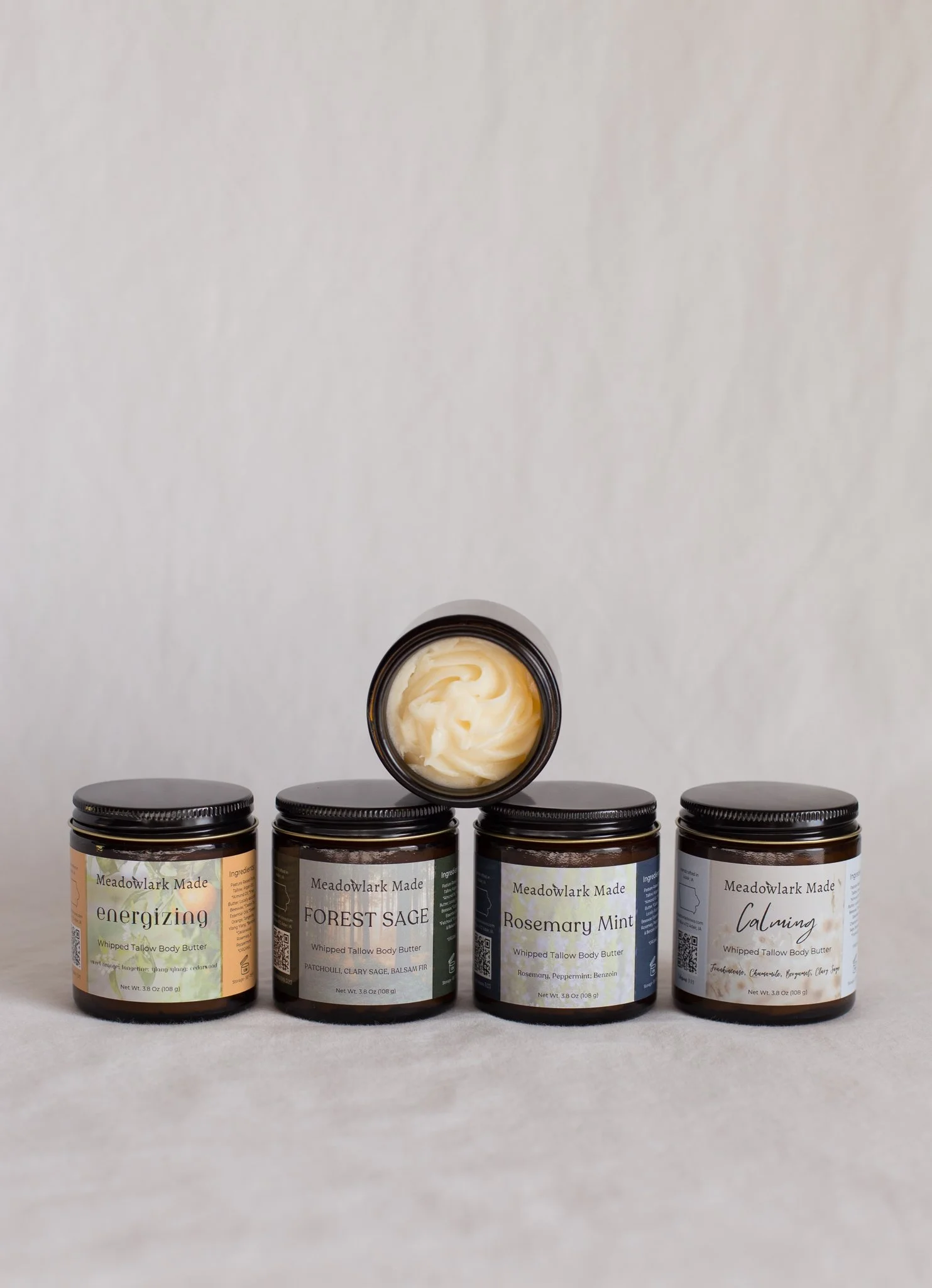Four jars of Meadowlark Made whipped body butter, with different scents: energizing, forest sage, rosemary mint, and calming. One jar is open, showing the nourishing pork tallow body butter inside.