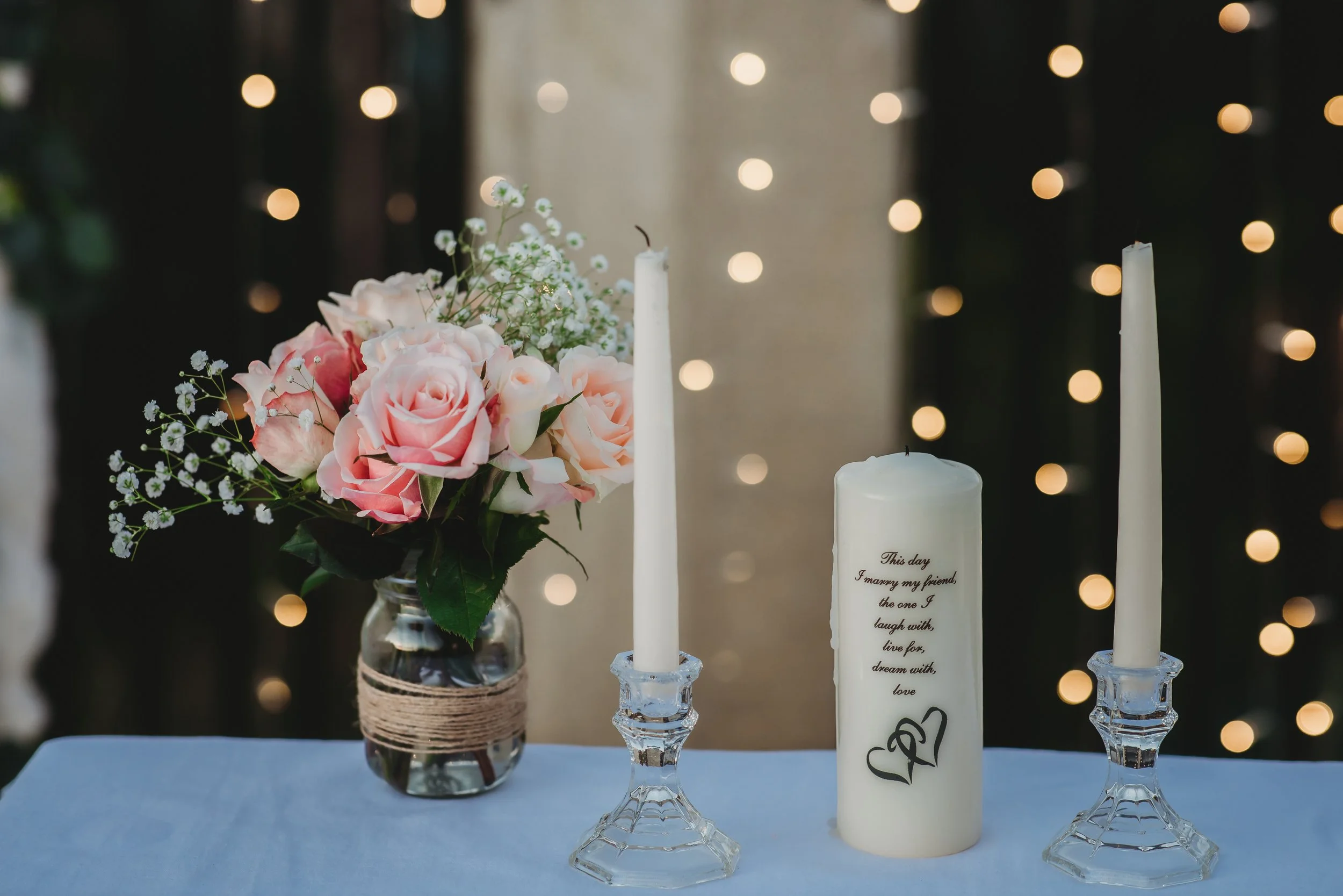 A floral arrangement of pink and white roses with baby's breath in a glass jar on a table. Two white taper candles are in glass candlestick holders, and a larger white pillar candle with black writing and heart design are in the middle. Blurred strin
