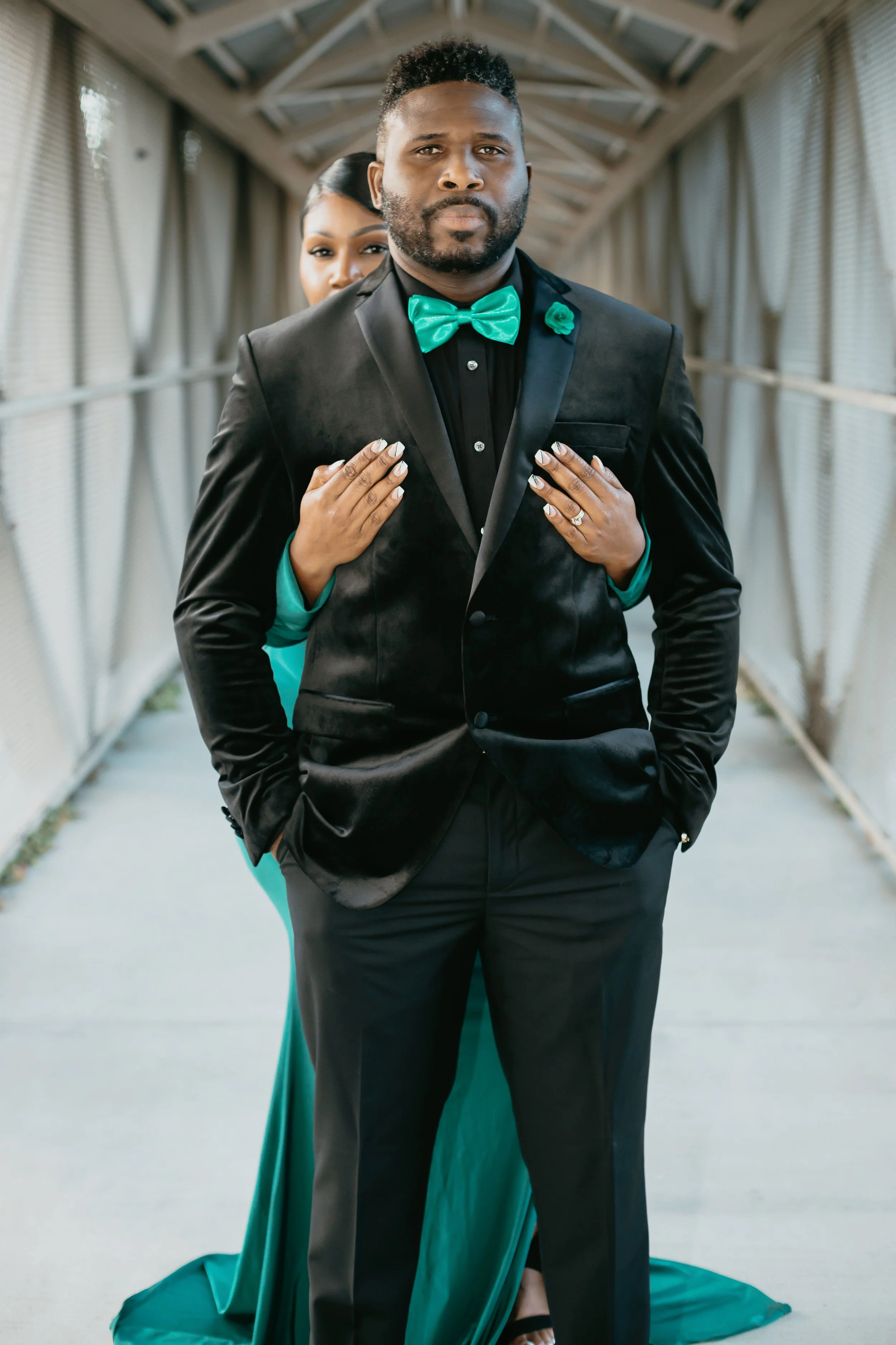 A man in a black tuxedo with a teal bow tie and boutonniere, standing confidently in a modern, covered walkway, with a woman in a teal gown standing behind him, both touching his shoulders.